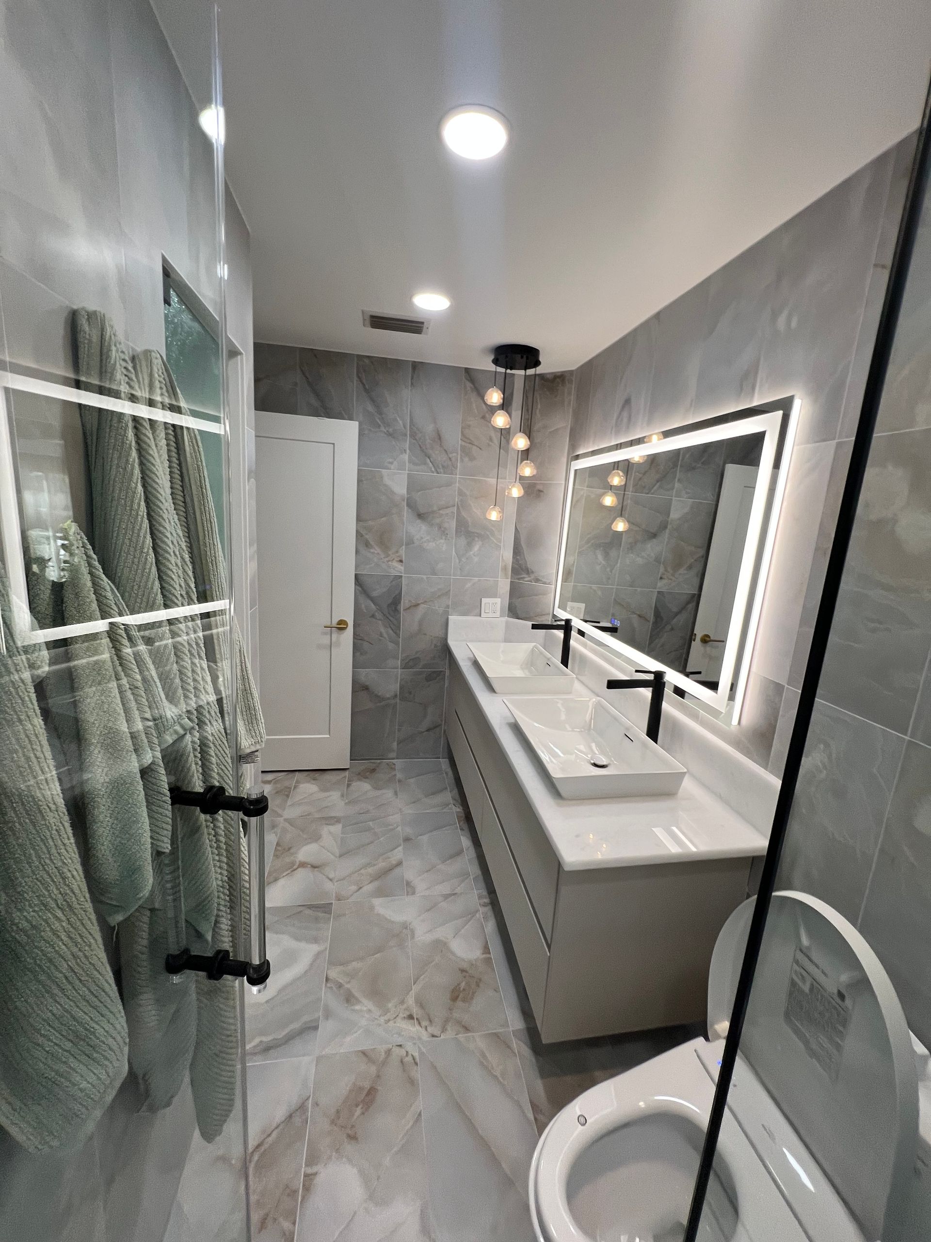 Modern bathroom with gray tile, double vanity, and toilet. Towels hang on a rack.