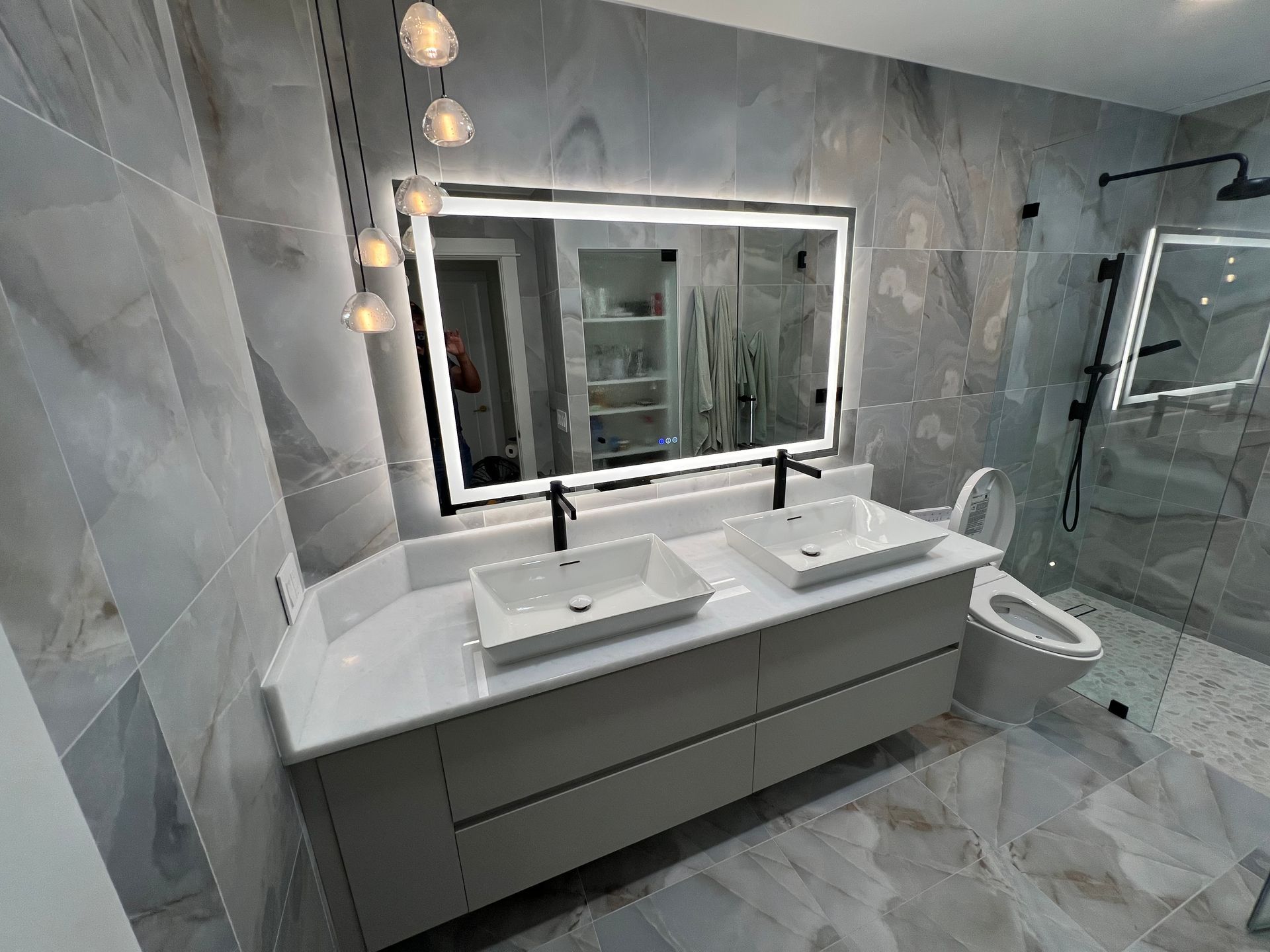 Modern bathroom with gray tile, double sinks, illuminated mirror, and glass shower.