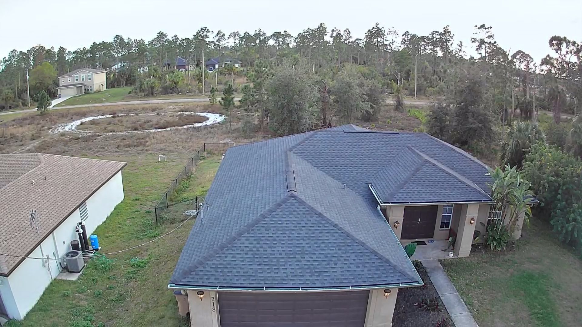 An aerial view of a house with a dark roof and a tan garage, set amidst trees and other houses.