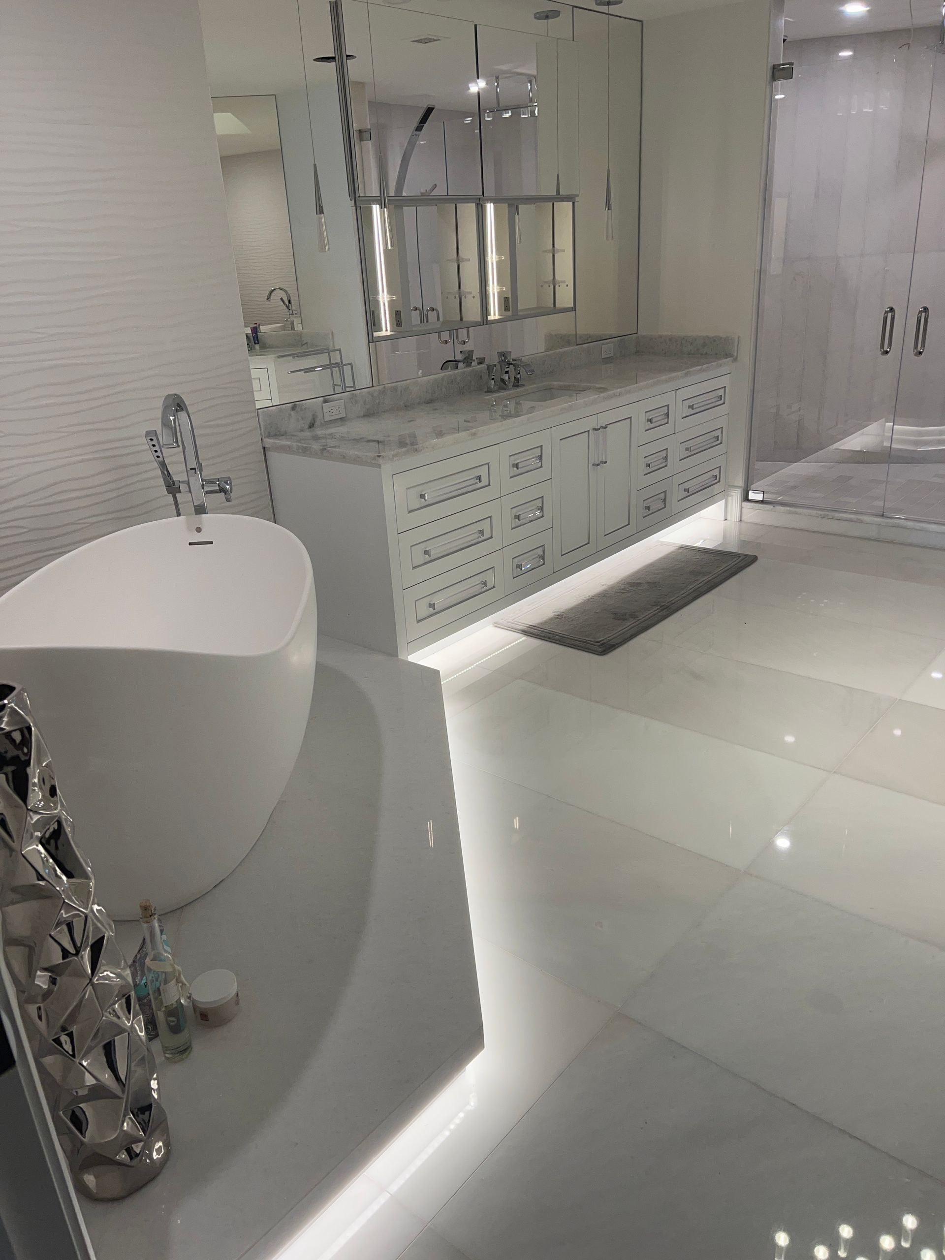 Modern all-white bathroom with a freestanding tub, vanity, and large mirror. Shiny white floor and ambient lighting.