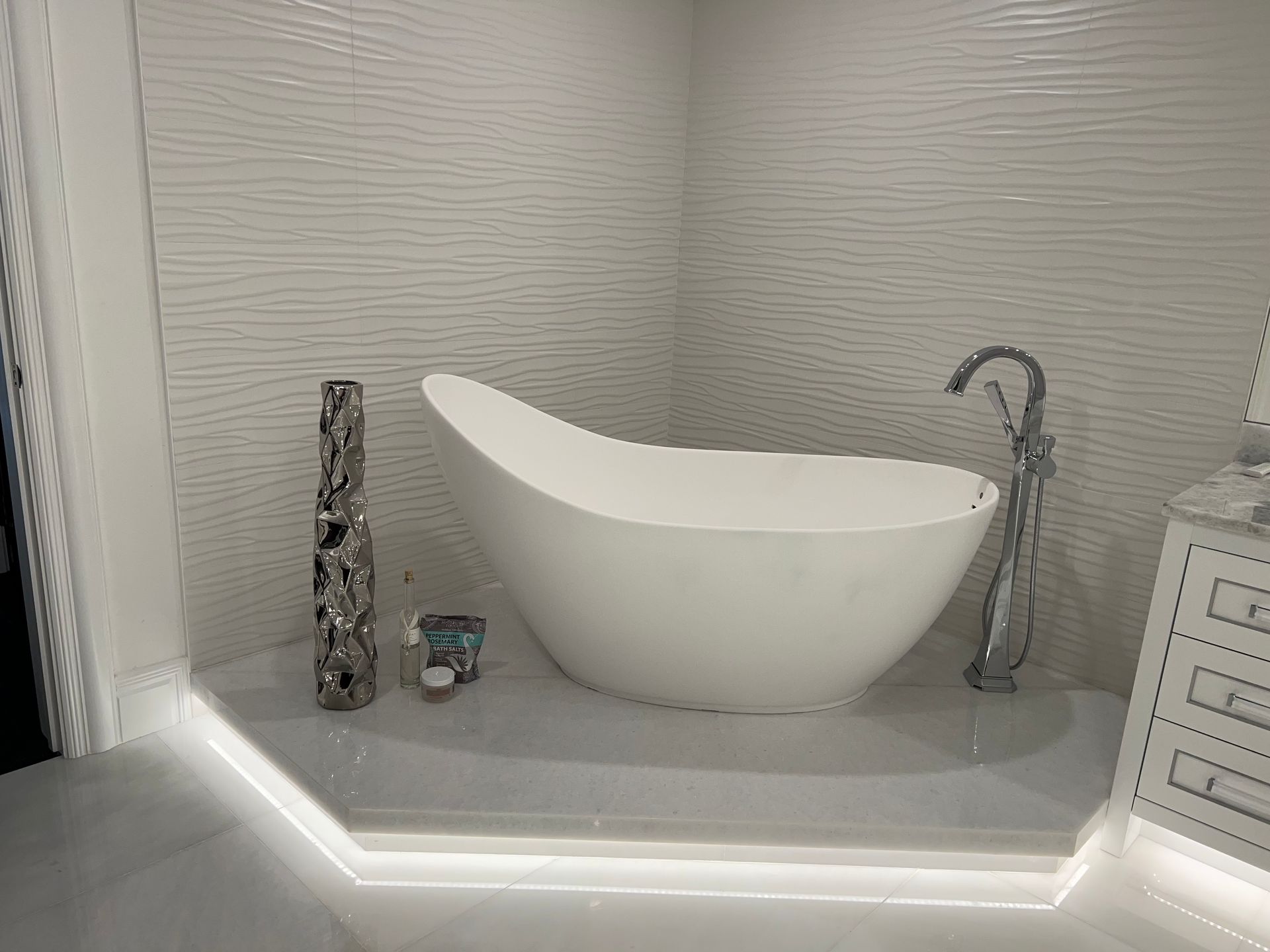 A white, modern bathtub in a bathroom with textured white walls. A decorative vase and candle are on the tub's ledge.