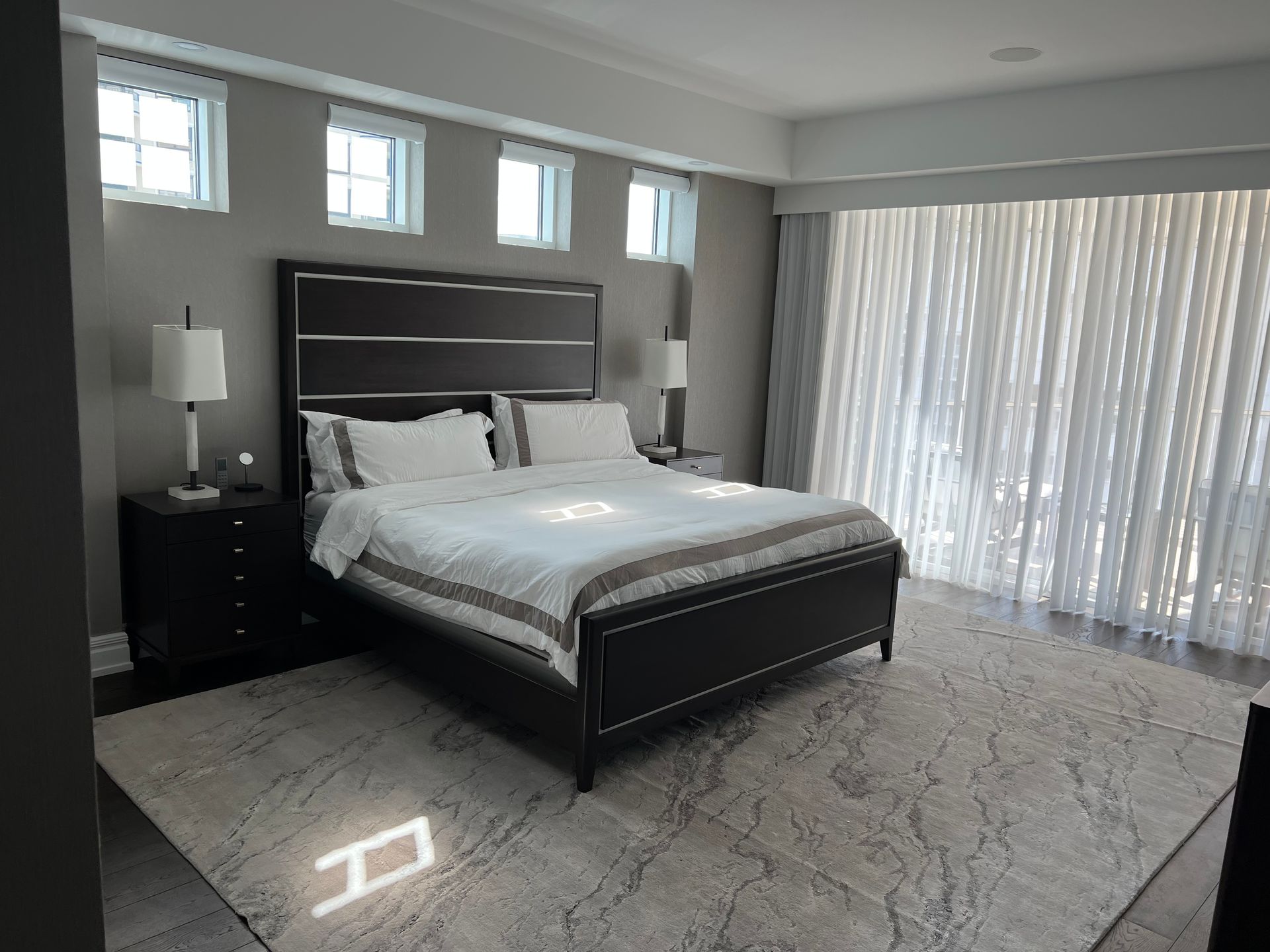 Bedroom with a dark bed, nightstands, rug, and sheer curtains; gray walls and windows.