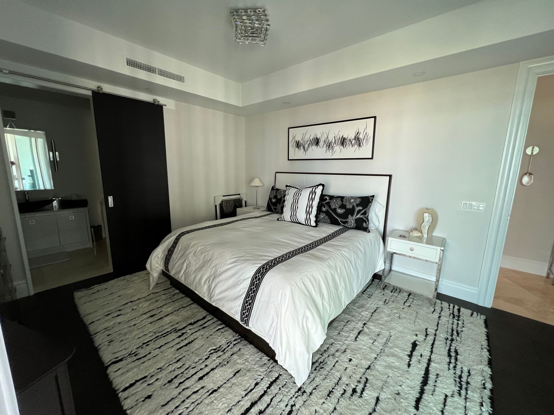 Bedroom with white bedding, black accents, and a black rug. Bathroom visible through a sliding door.
