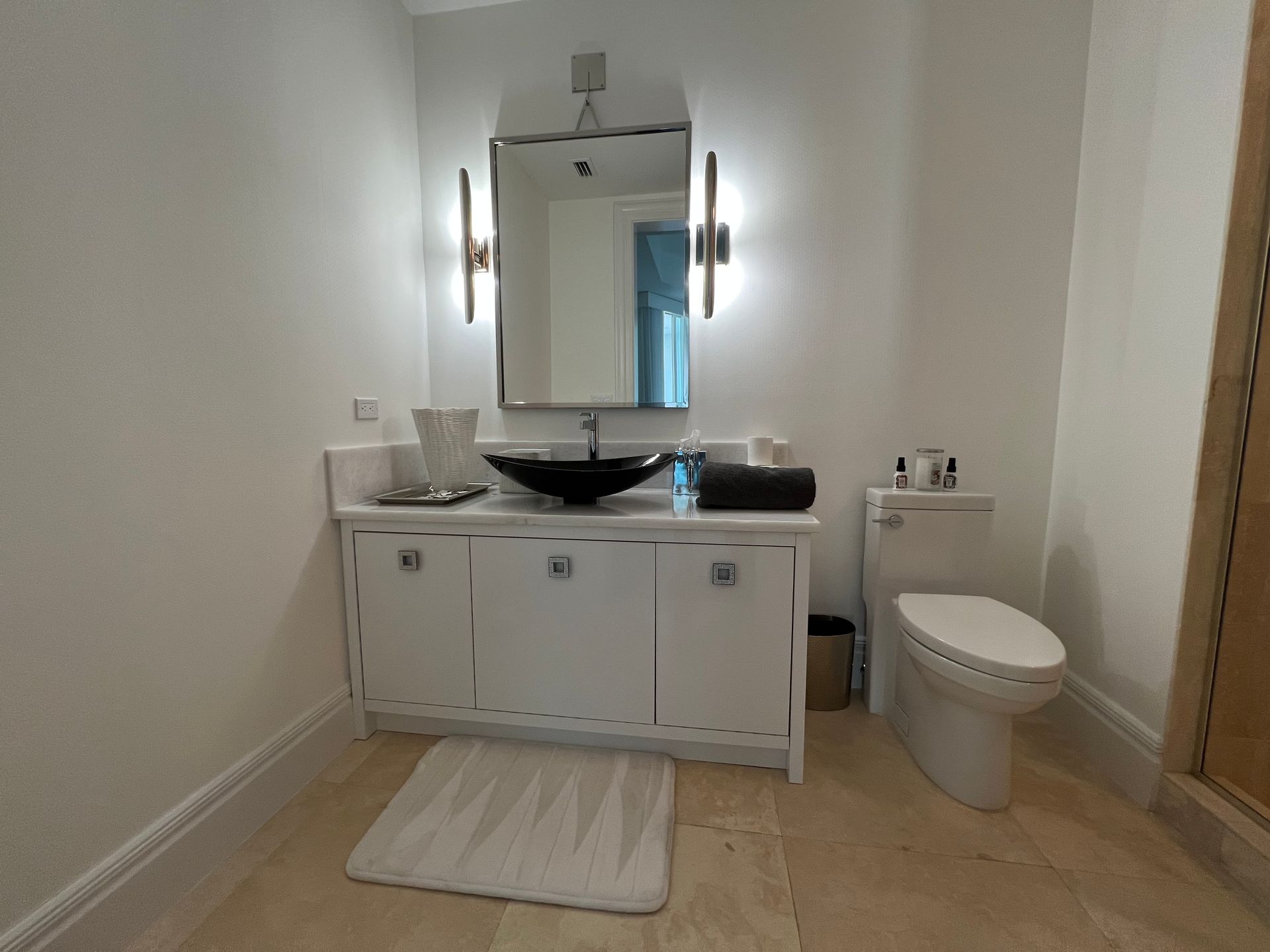 White bathroom with vanity, mirror, toilet, and cream rug.
