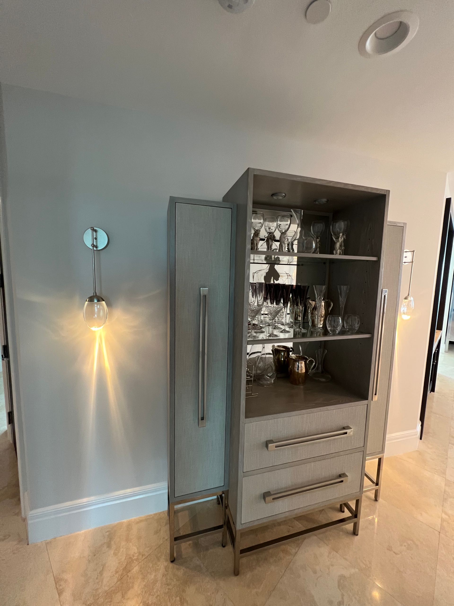 Gray bar cabinet with glass shelves and drawers, next to a wall sconce, set on a light-colored floor.
