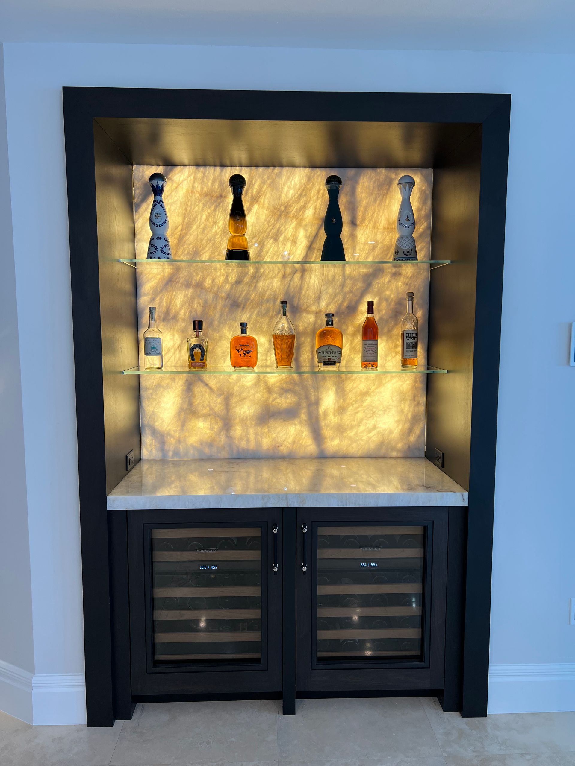 A custom bar with backlit display shelves holding liquor bottles, above a cabinet with two wine coolers.