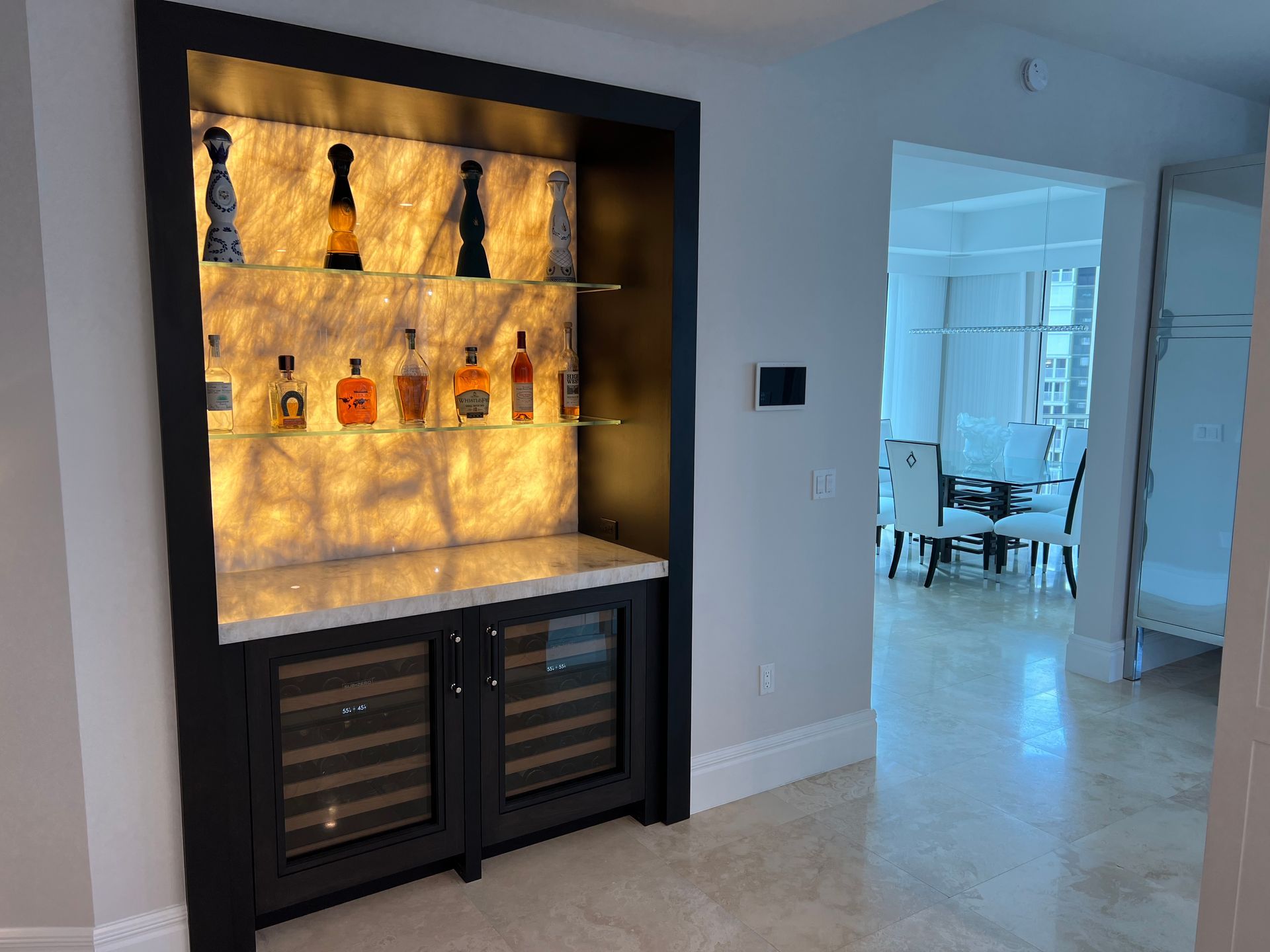 Built-in bar with backlit liquor display, wine fridge, and a glimpse of a dining area.