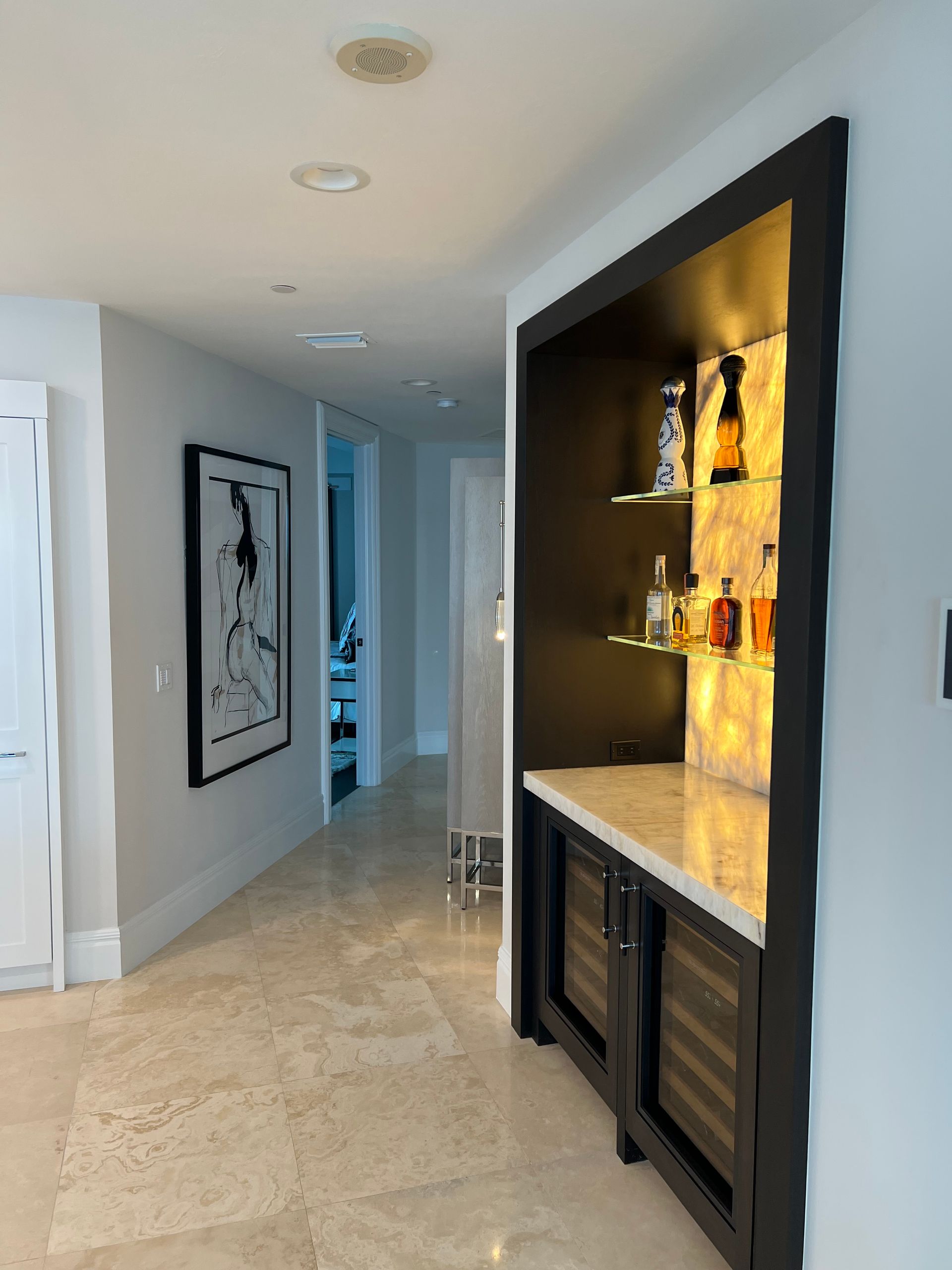 Hallway with built-in bar, including wine fridge, shelves, and backlit onyx. Light-colored walls and flooring.