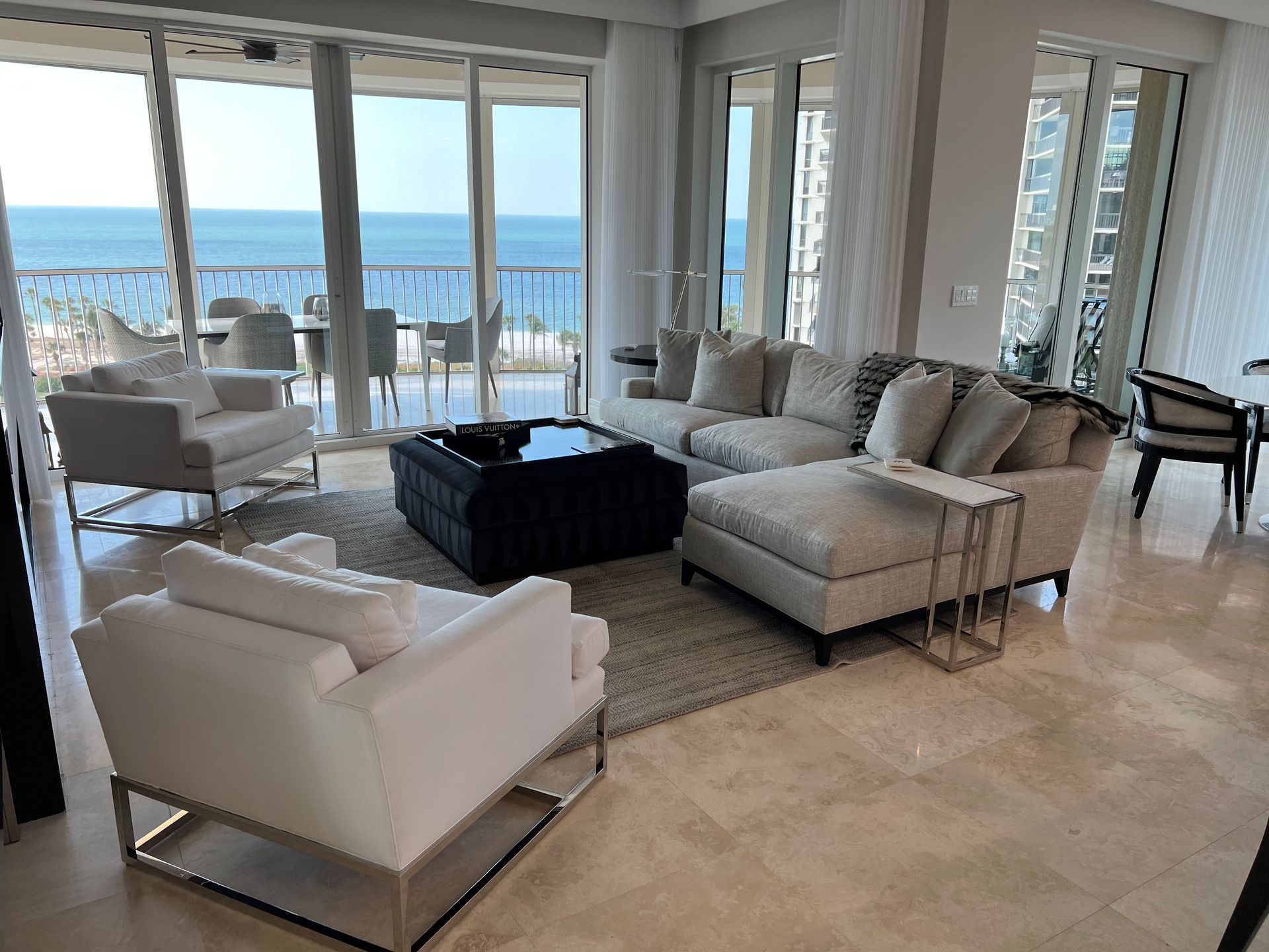 Living room with ocean view: white furniture, large windows, blue water, and sunny outdoor balcony.