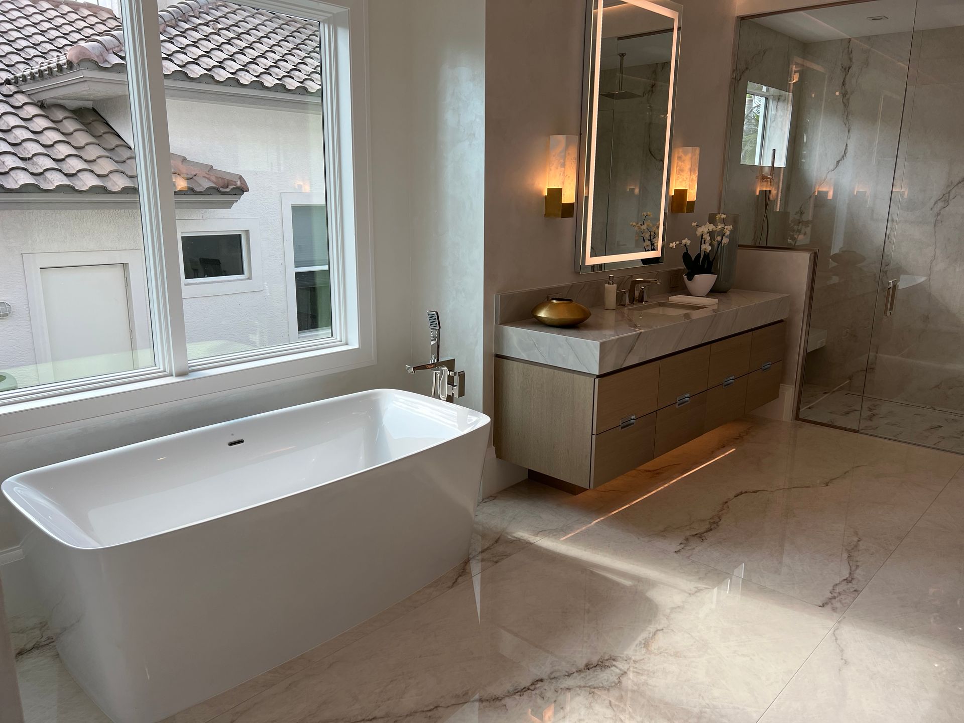 Modern bathroom with a white tub, marble floors, and a vanity, bathed in sunlight.