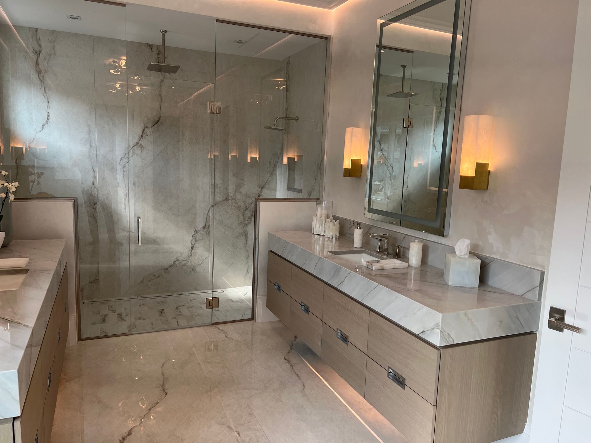 Modern bathroom with marble, wood vanity, glass shower, and gold sconces.