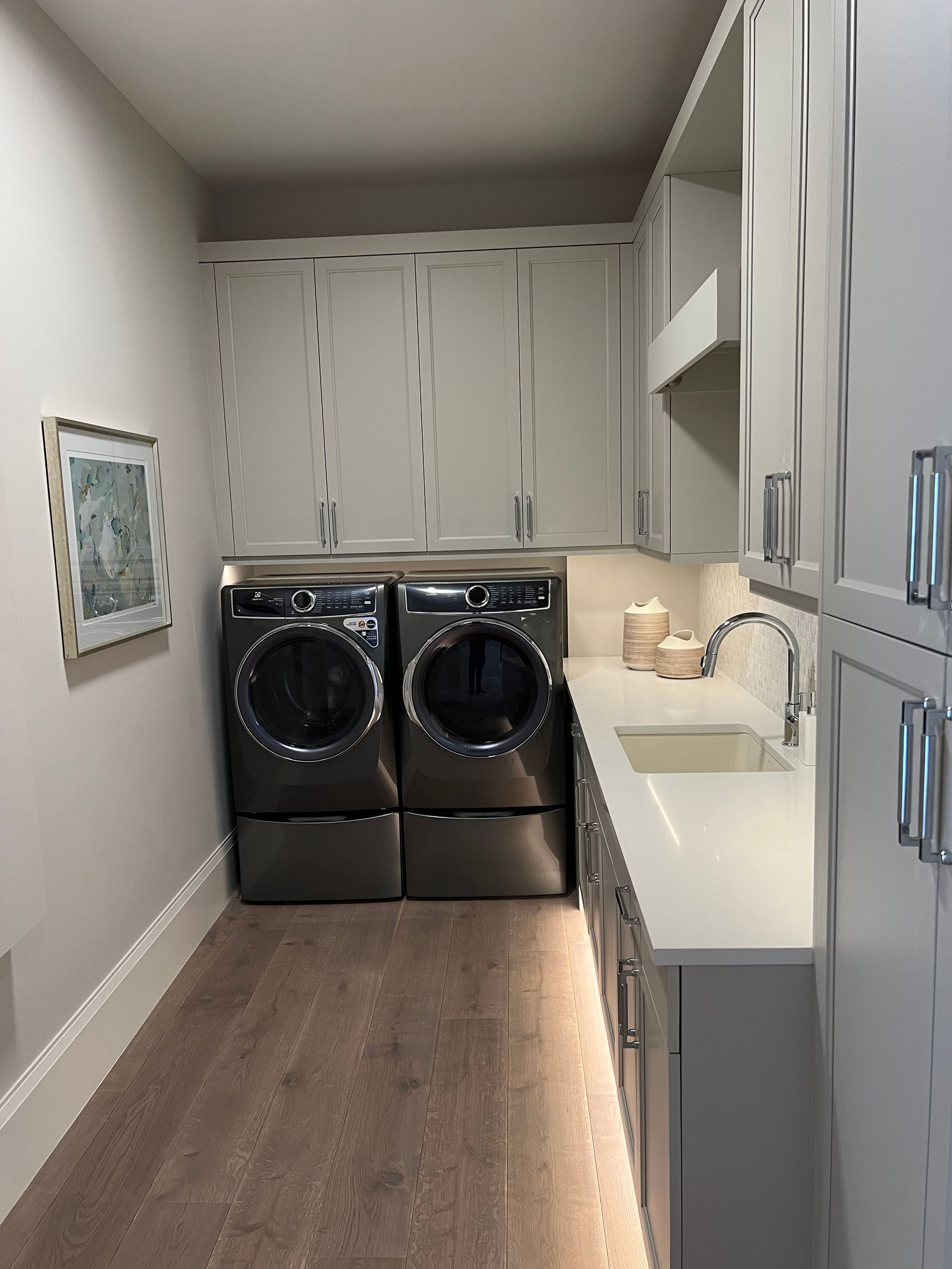 Laundry room with washer/dryer, cabinets, sink, and light wood flooring. Light gray and white tones.