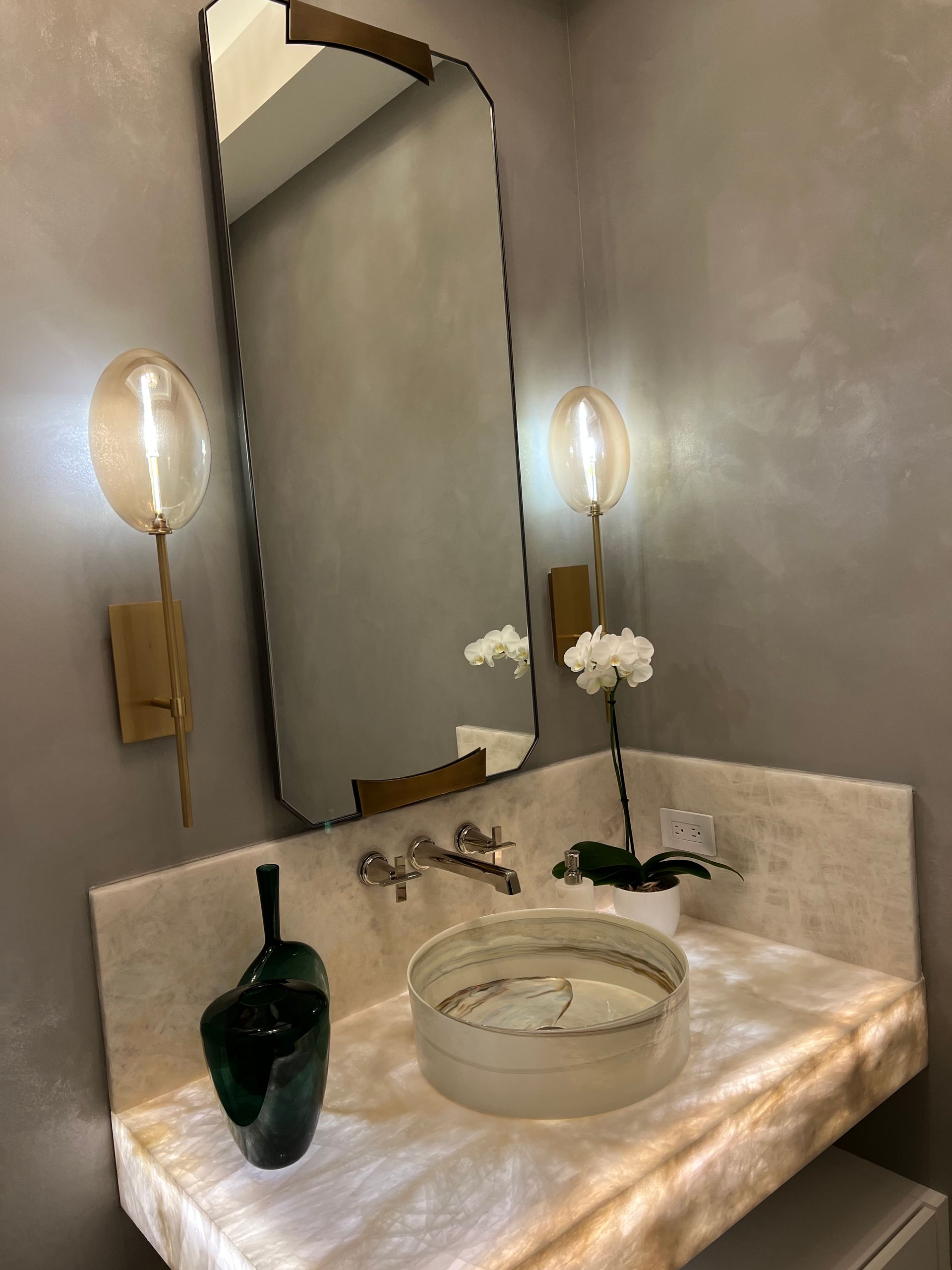 Bathroom with a backlit marble vanity, curved mirror, sconces, and vessel sink with white orchids.