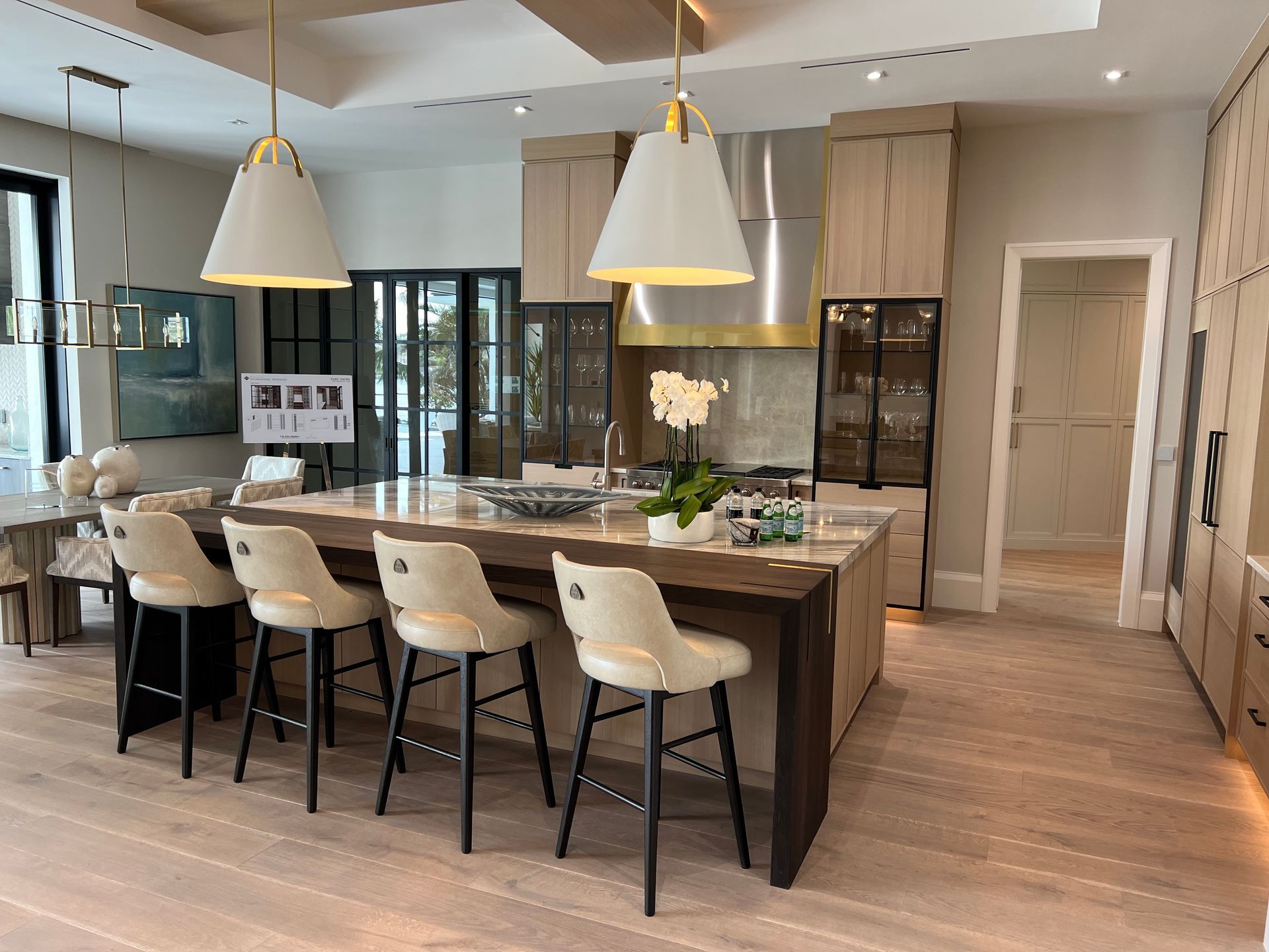 A modern kitchen with a large island, bar stools, and pendant lights. Light wood floors and cabinets.