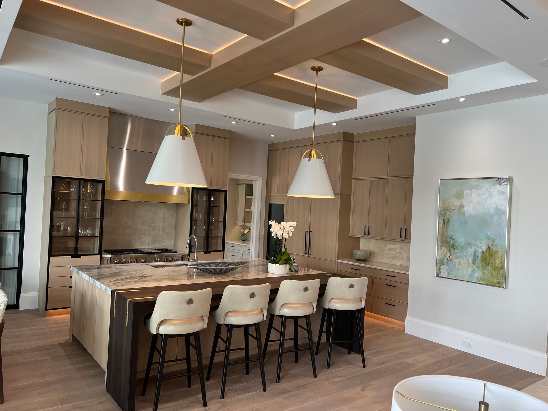 A modern kitchen with light wood cabinets, island, and pendant lights, featuring seating and a painting.