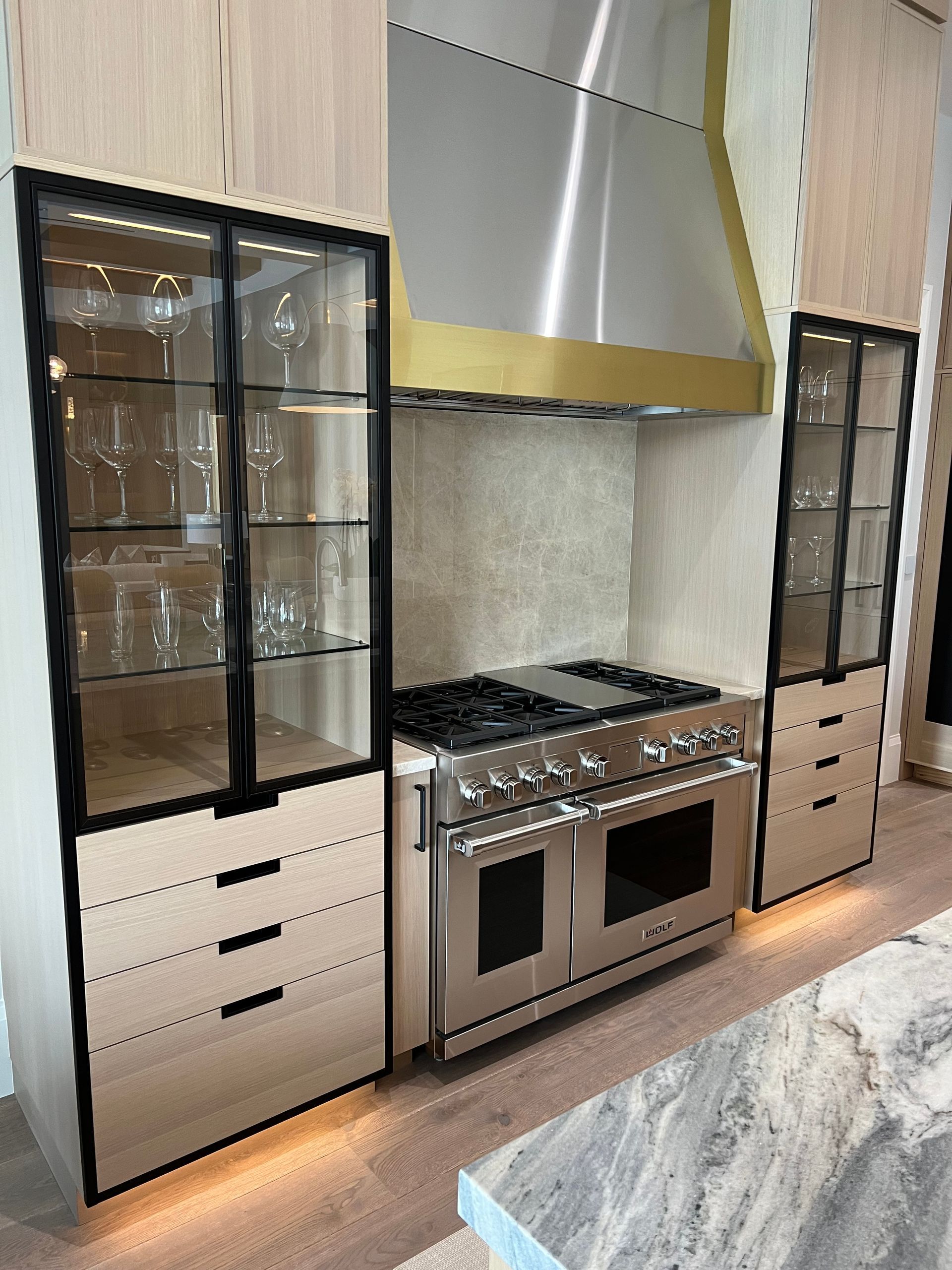 Modern kitchen with stainless steel range, brass range hood, and light wood cabinets with black trim and glass doors.
