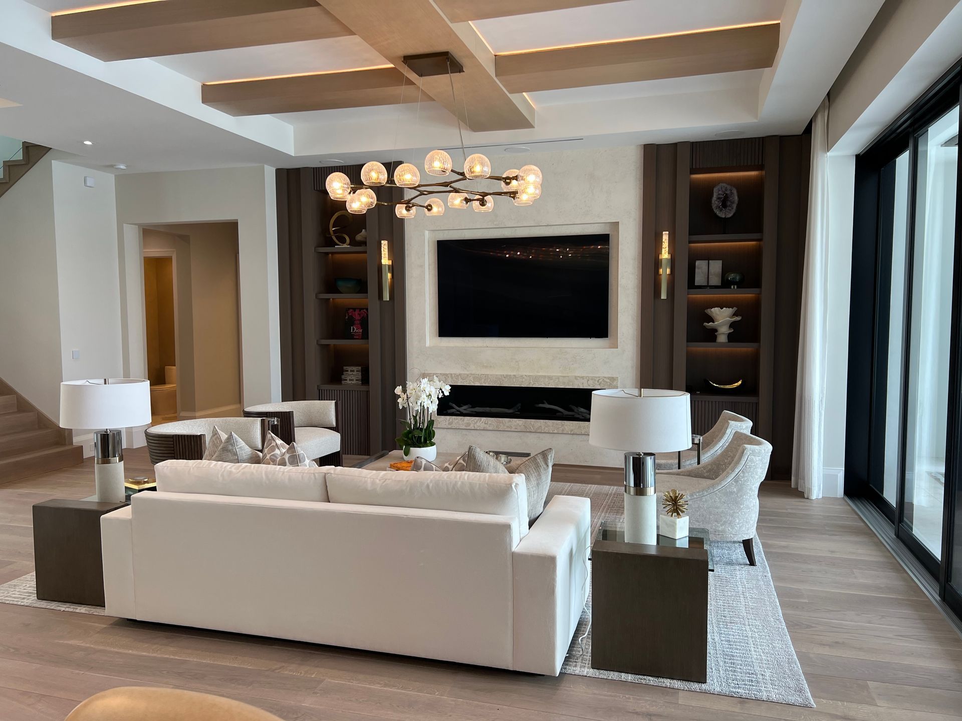Living room: white sofa, fireplace, TV, shelves, chandelier, wood beams, light walls, chairs, side tables, large windows.