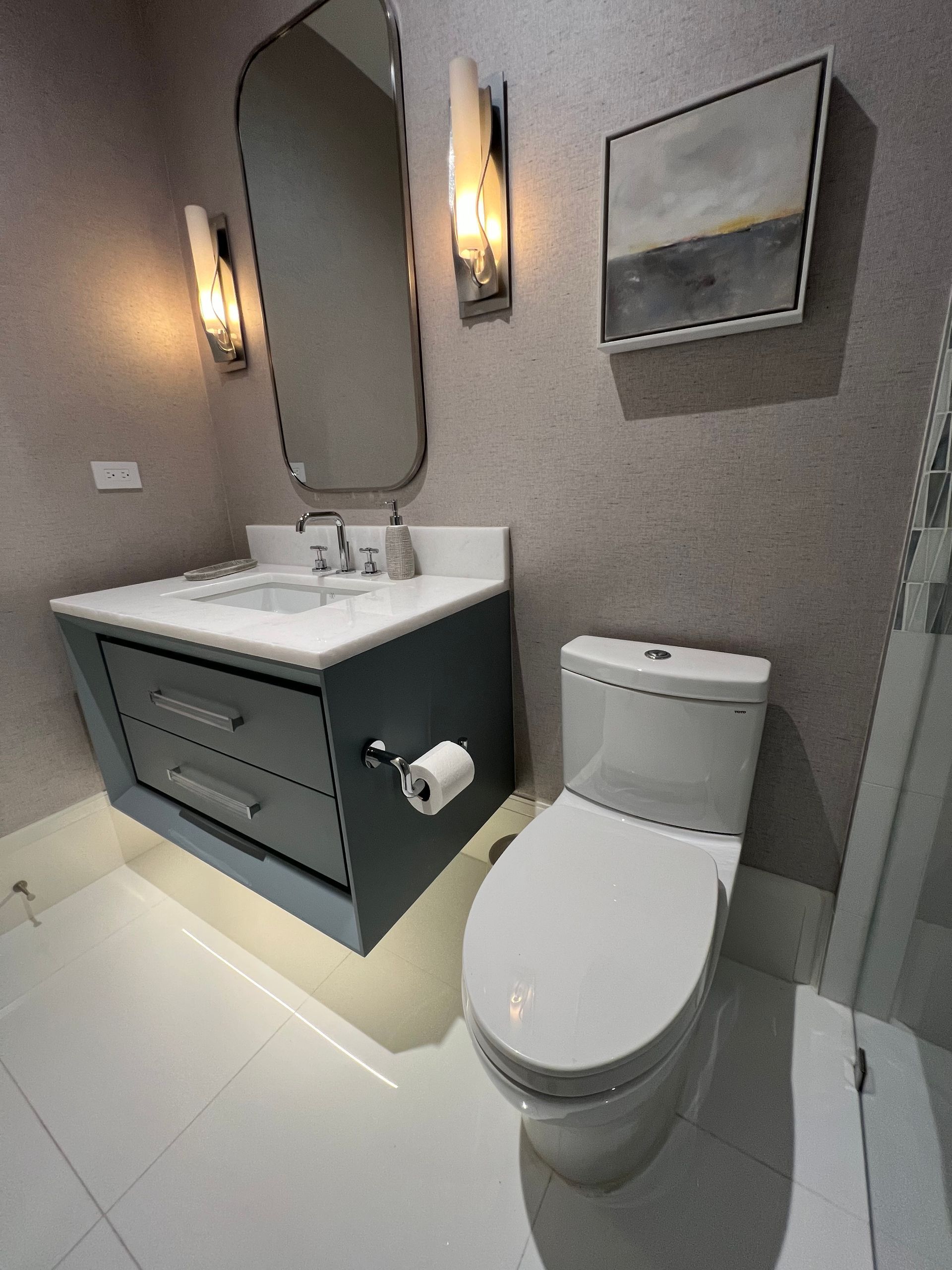 Powder room with floating blue vanity, toilet, mirror, art, and wall sconces on gray textured wall.