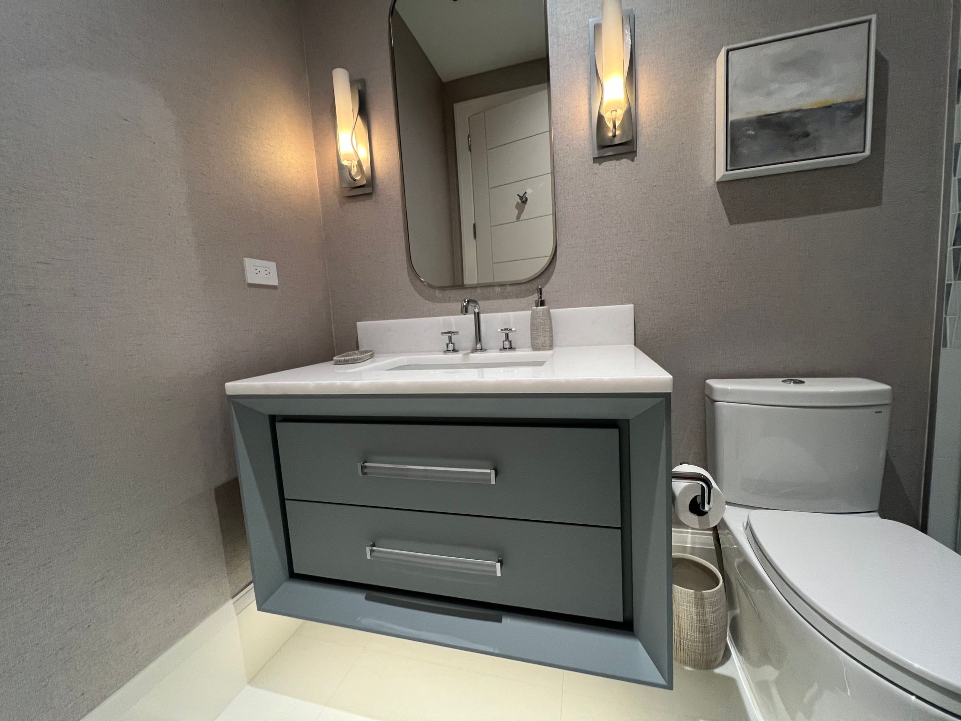 Small modern bathroom with a blue vanity, mirror, sconces, and toilet. Grey textured walls.
