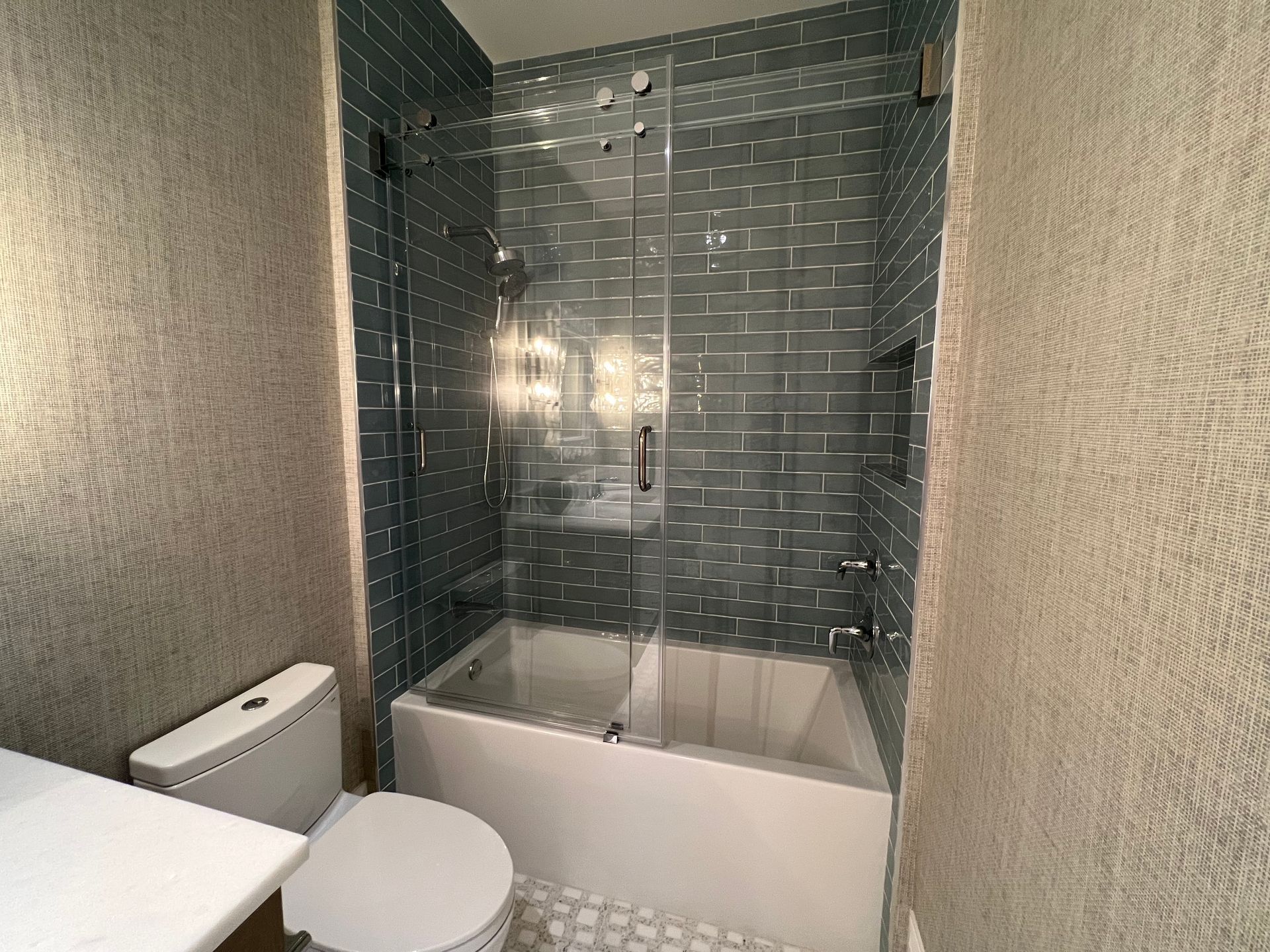 Small bathroom with white tub, toilet, shower, glass doors, blue tile, and beige patterned walls.