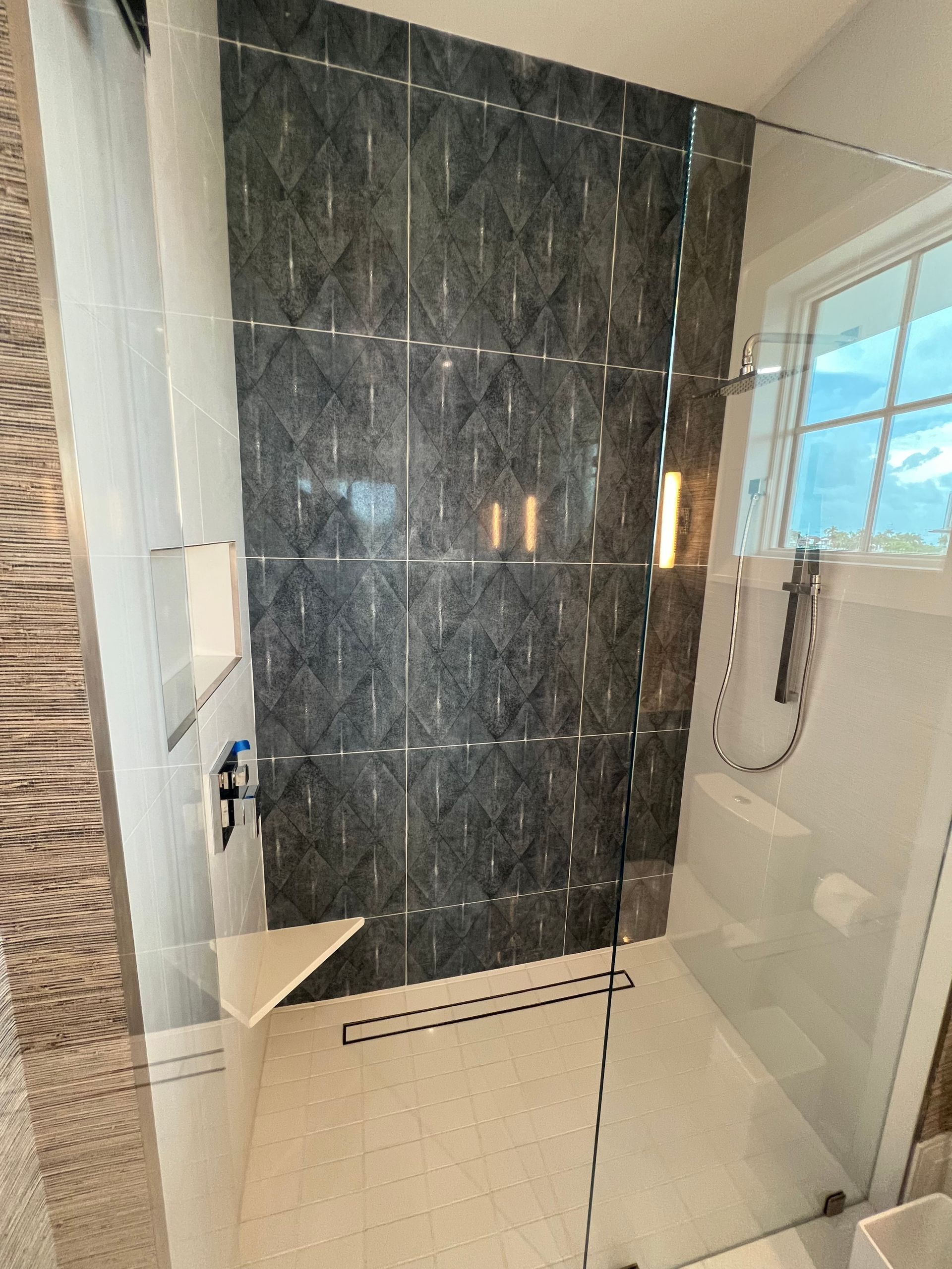 Modern bathroom shower with gray patterned tile walls, glass door, and white tiled floor.