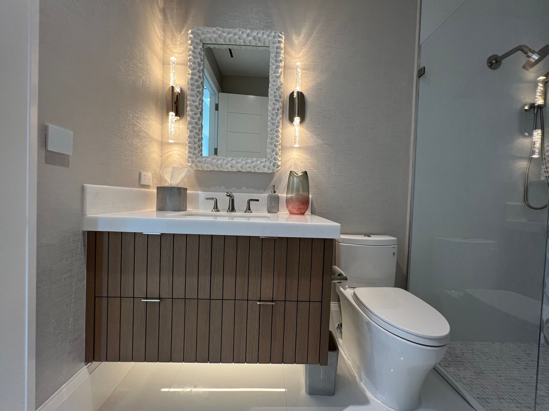 Modern bathroom with vanity, toilet, and shower. Brown textured cabinet, mirror with lights, white fixtures.