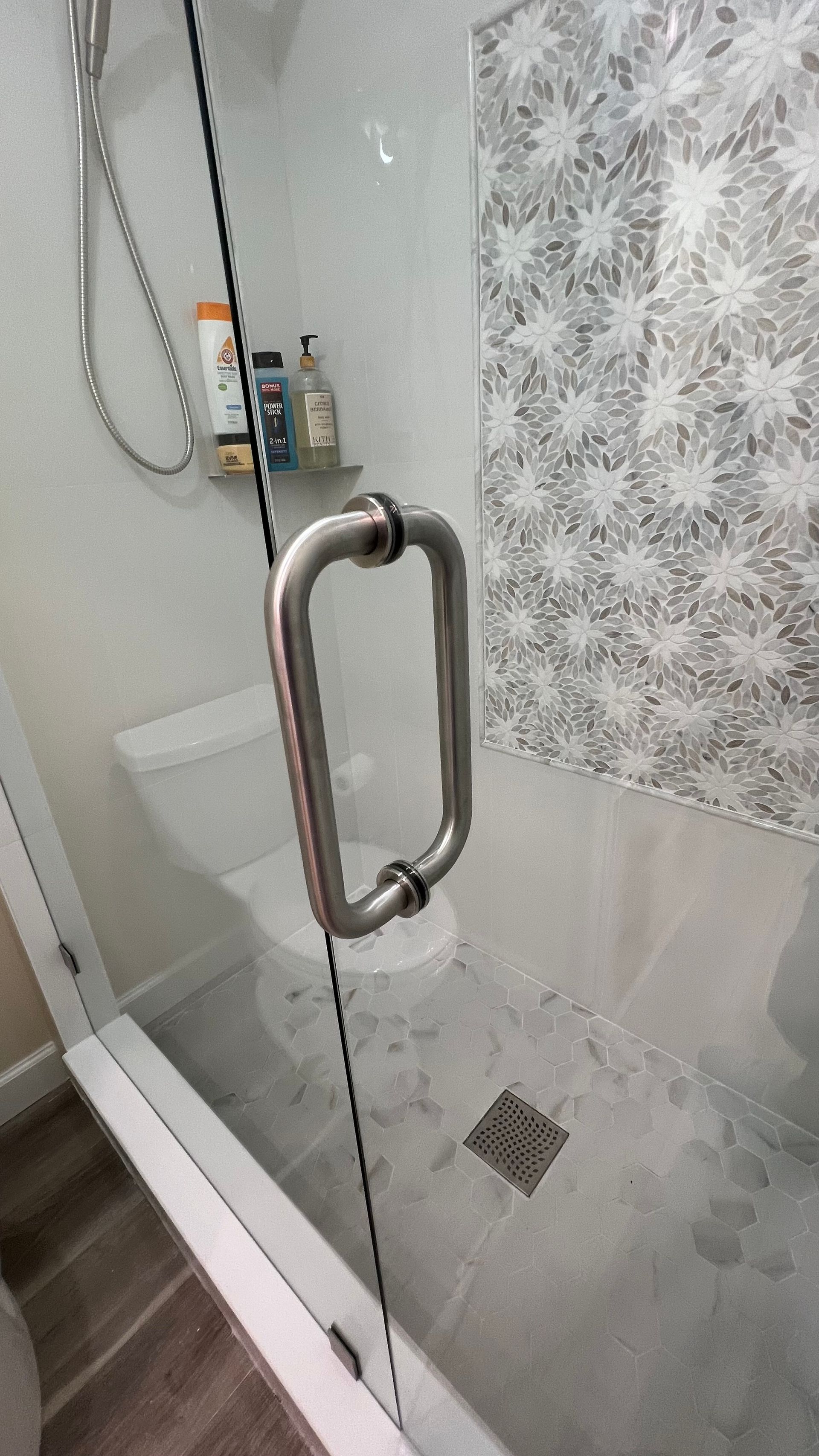 Shower with glass door, stainless steel handle, tiled accent wall, toiletries.