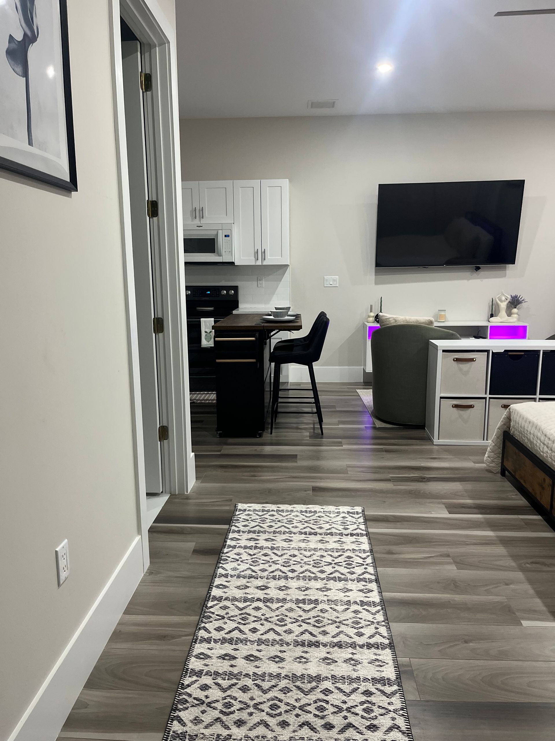 Narrow hallway with a patterned rug leading to a kitchen and living area; a TV is mounted on the wall.