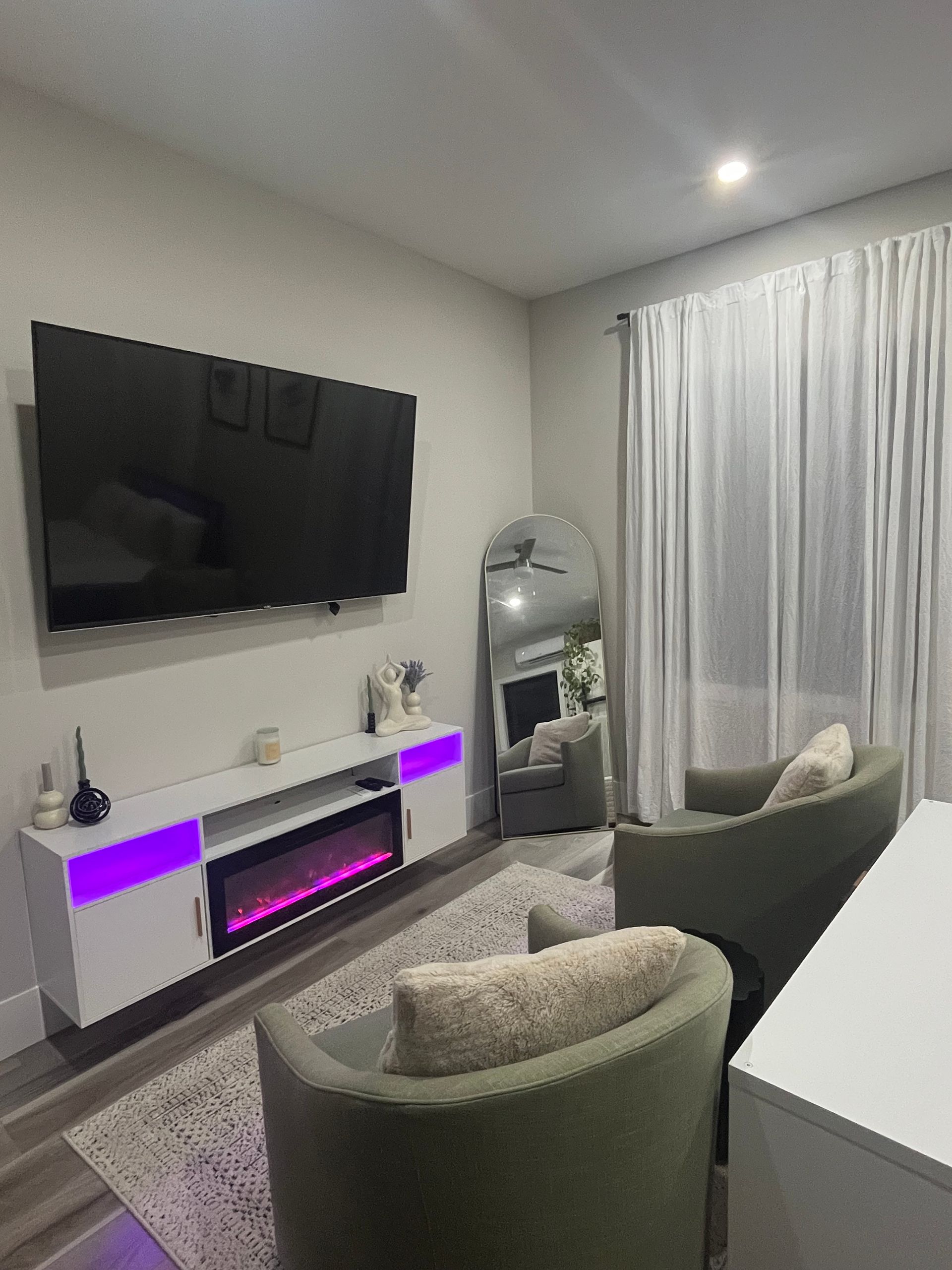 Living room with mounted TV, fireplace, green chairs, large mirror, and sheer curtains.