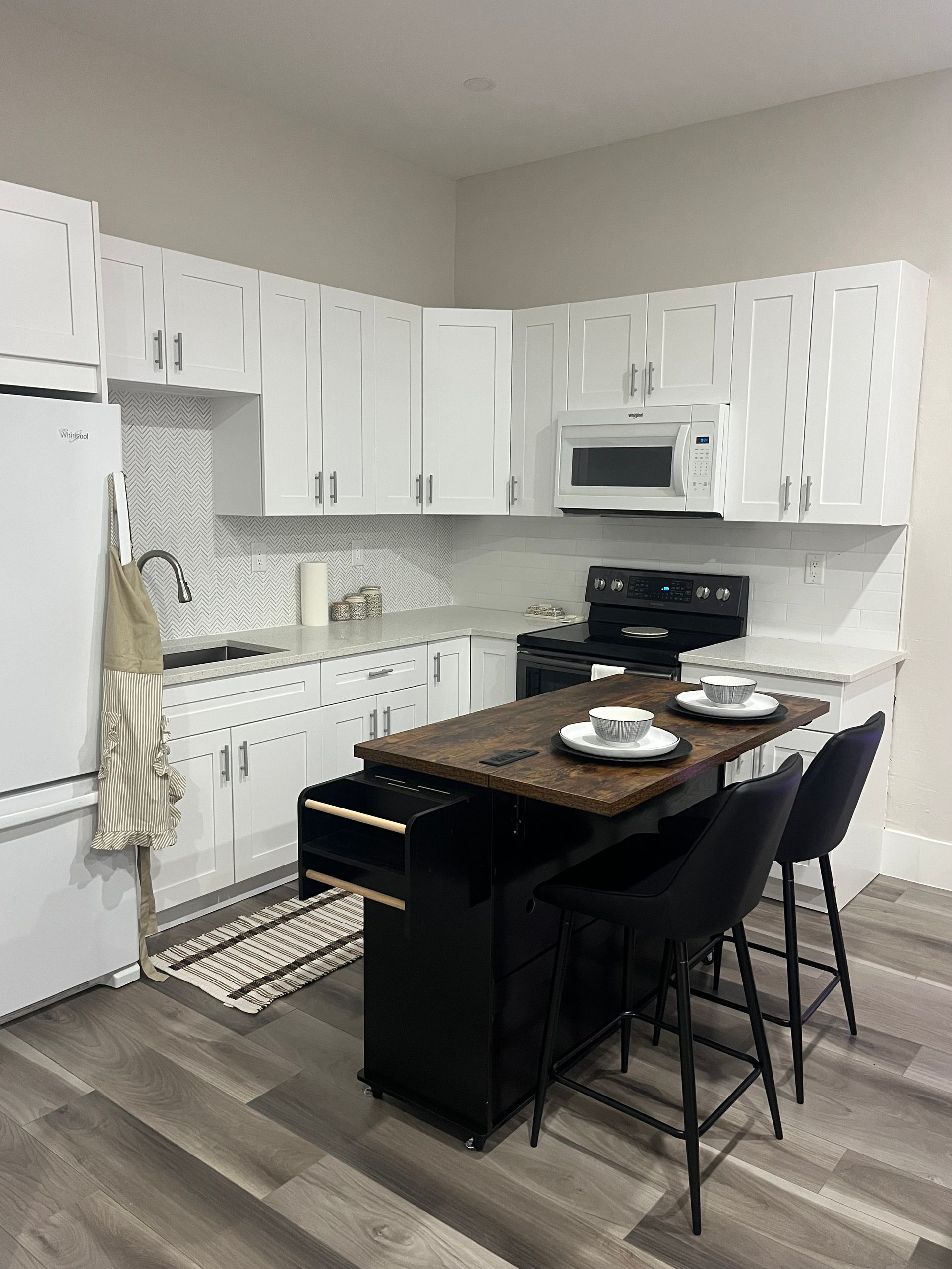 Small white kitchen with black bar and three stools. Wooden countertop, white cabinets, and gray flooring.