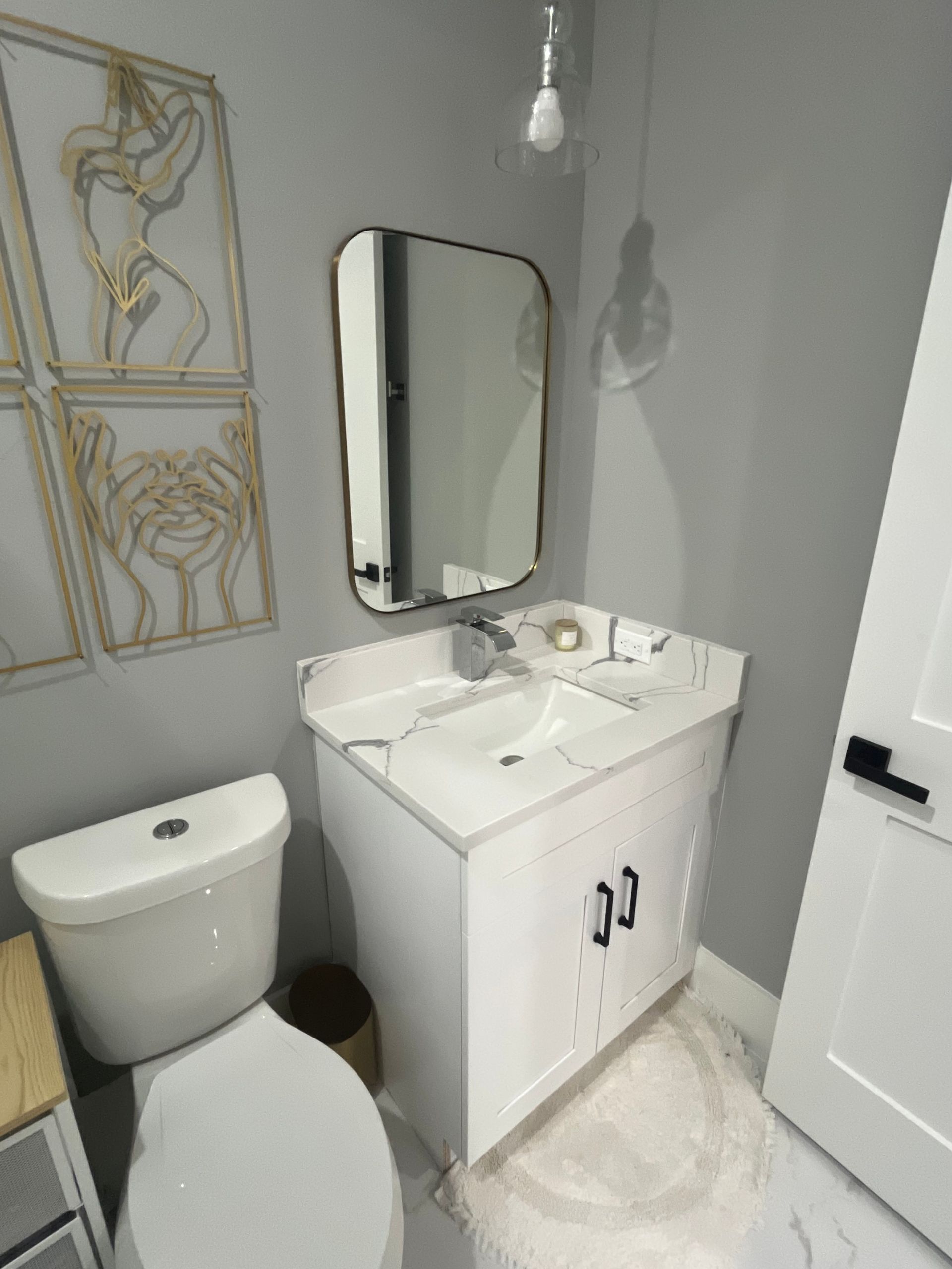 Small bathroom with toilet, vanity, mirror, and wall art. Gray walls, white fixtures, and decorative pieces.