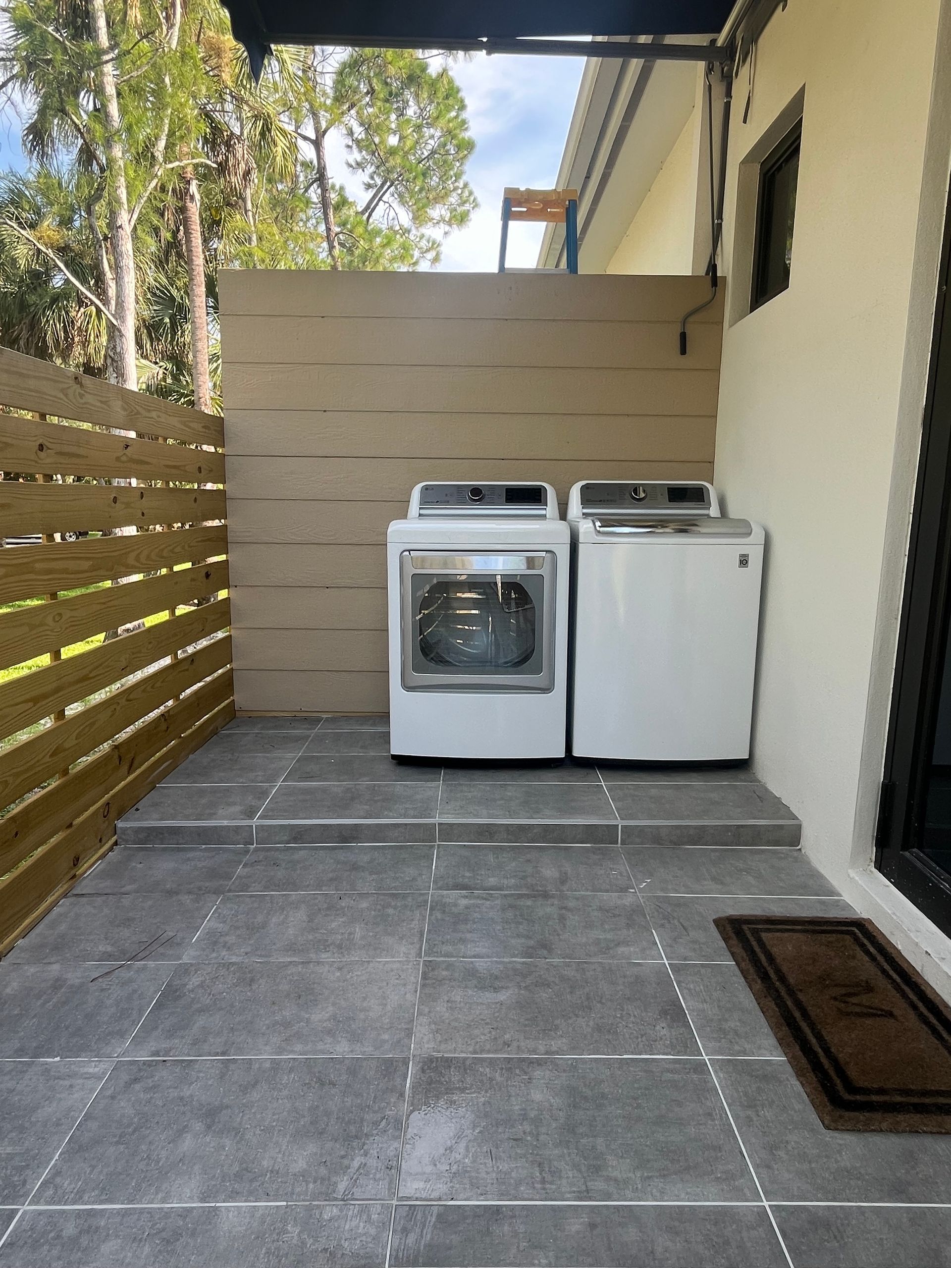 Outdoor laundry area with white washer and dryer, gray tile, and wooden fence.