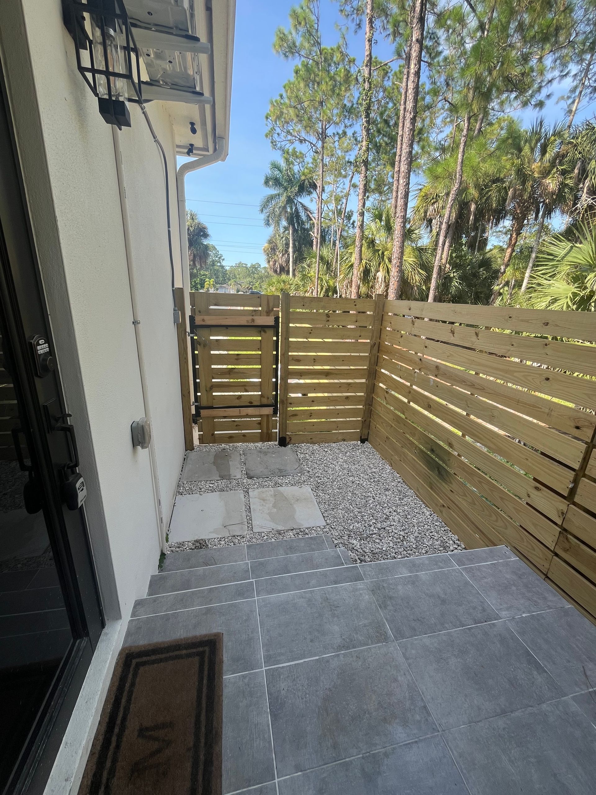 Exterior patio with gray tiles, steps, gravel, wooden fence and gate, and trees.