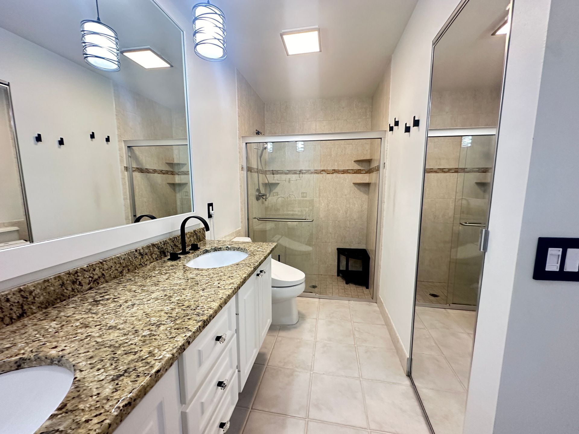 Bathroom with double sinks, granite countertop, glass shower, white cabinets, and light fixtures.