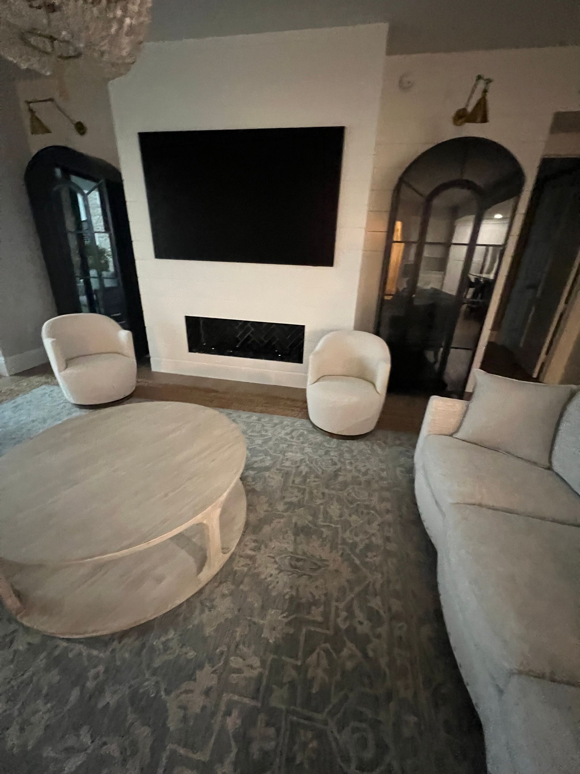 Living room with a TV above a fireplace, two white chairs, a sofa, and a round coffee table on a patterned rug.