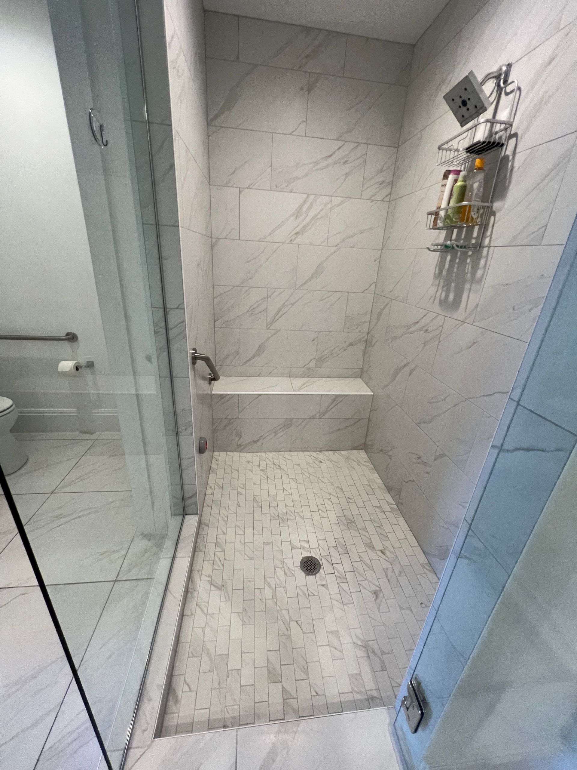 Marble-tiled shower with glass door, bench, and handheld shower head. Accessible bathroom with grab bar.