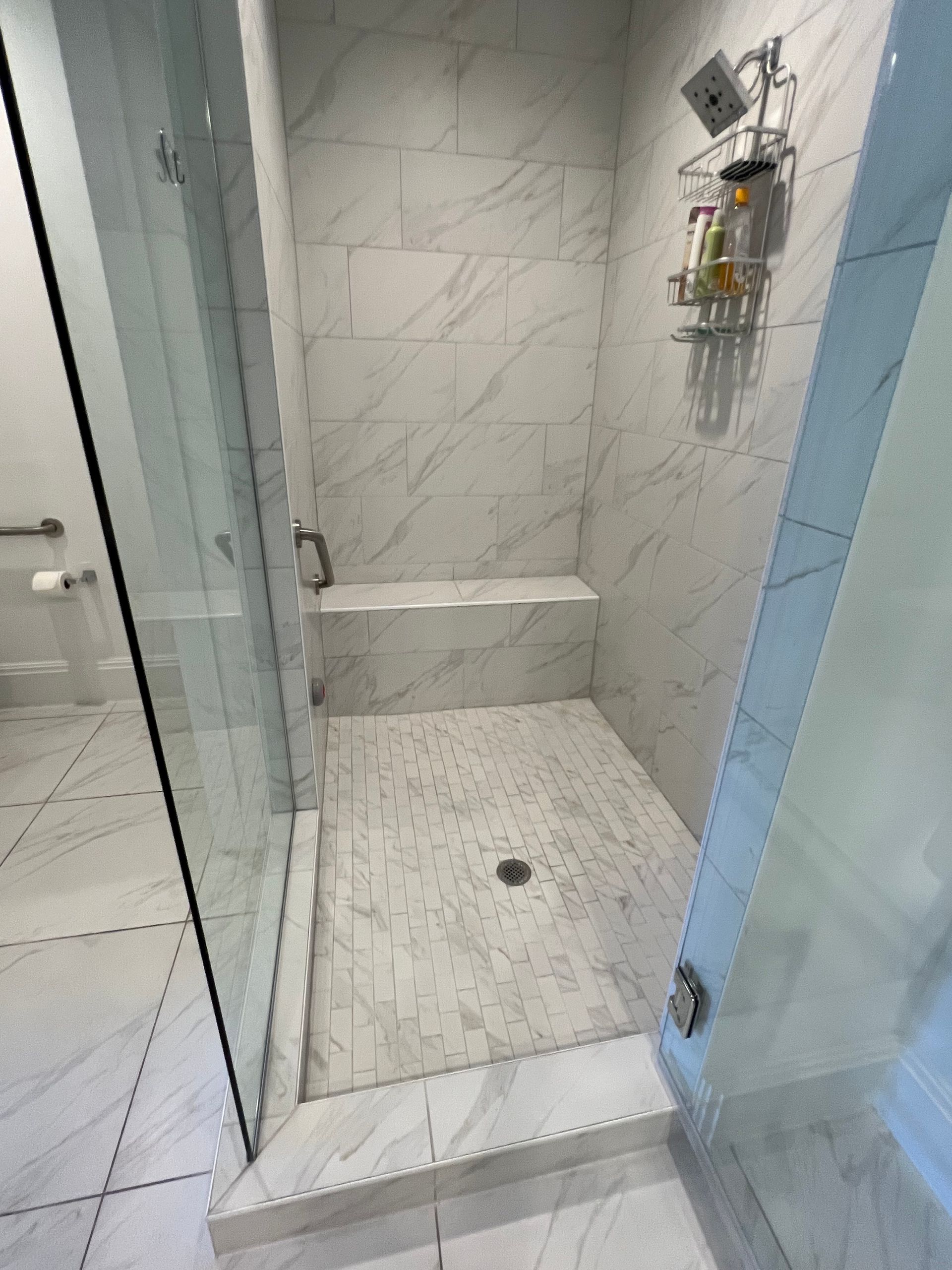 Walk-in shower with white marble tile, built-in bench, glass doors. Showerhead and toiletries visible.