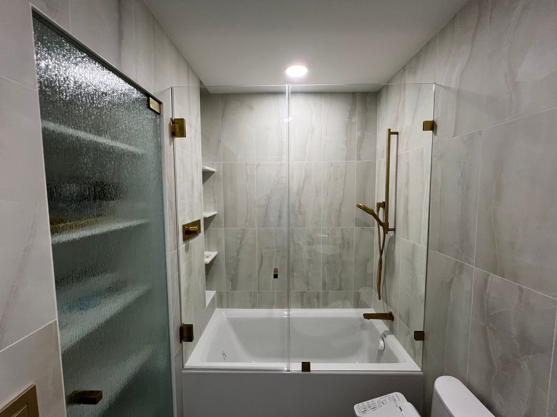 Modern bathroom with a white tub, glass shower doors, and gray tiled walls.