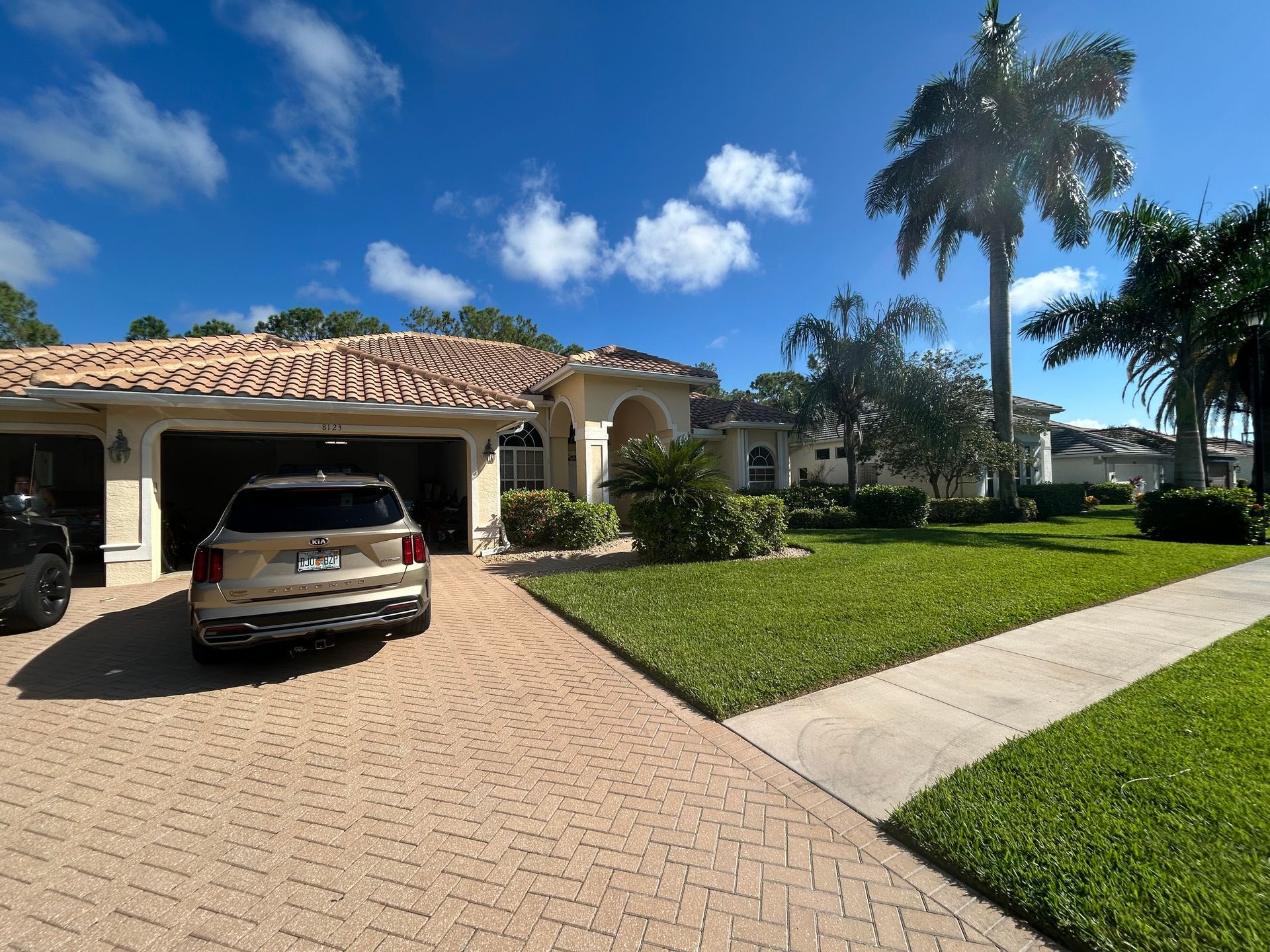 Beige house with tiled roof, SUV in driveway, green lawn, palm trees, and blue sky.