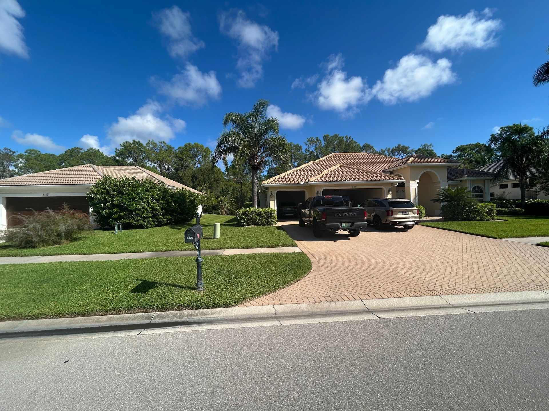 Suburban house with red-tiled roof, green lawn, brick driveway, cars parked in the garage, and bright blue sky.