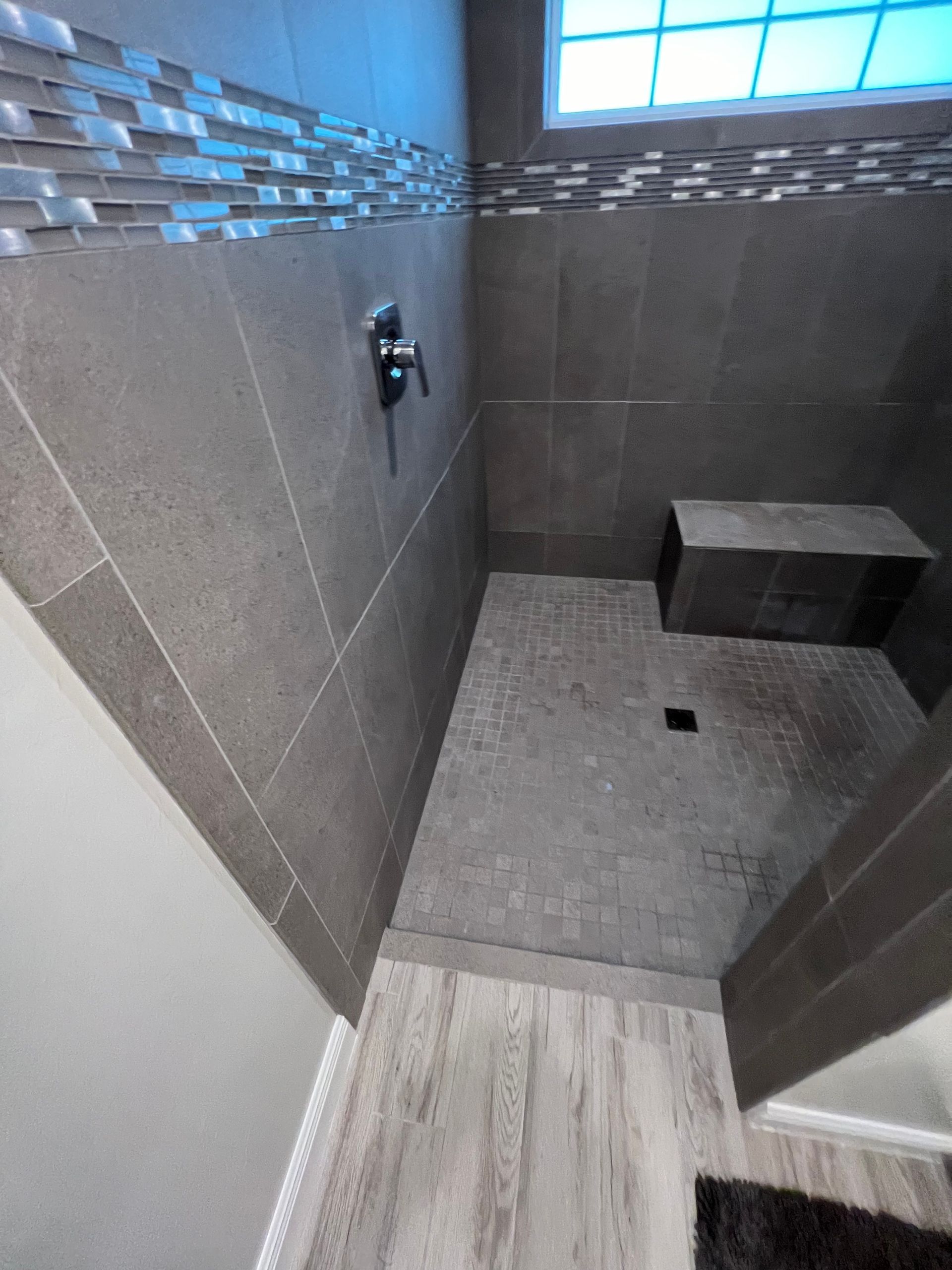 Gray tiled shower with a bench, silver fixtures, and a small window with blue glass.