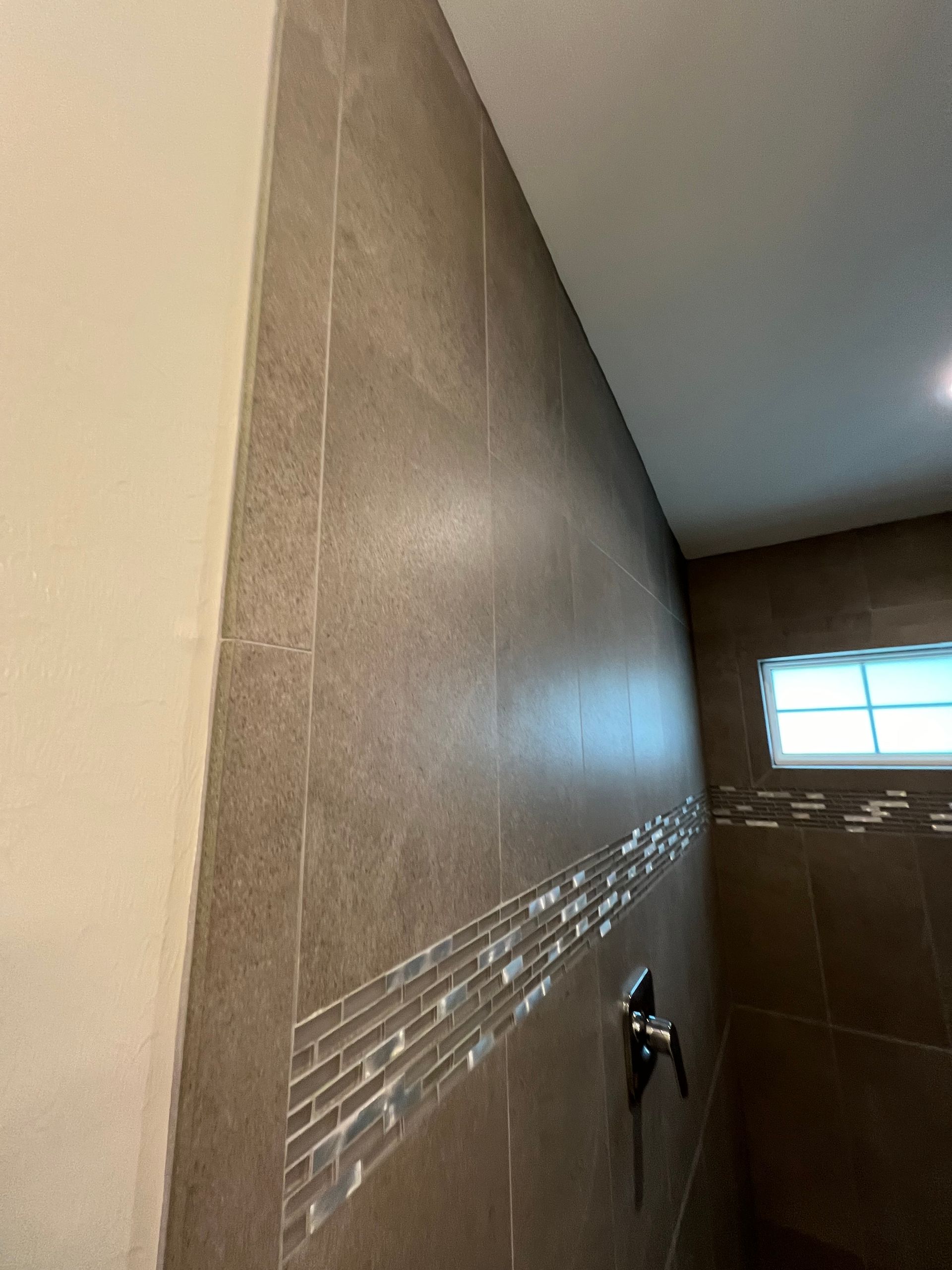 Brown tiled shower wall with a glass tile accent, a shower handle, and a small window.