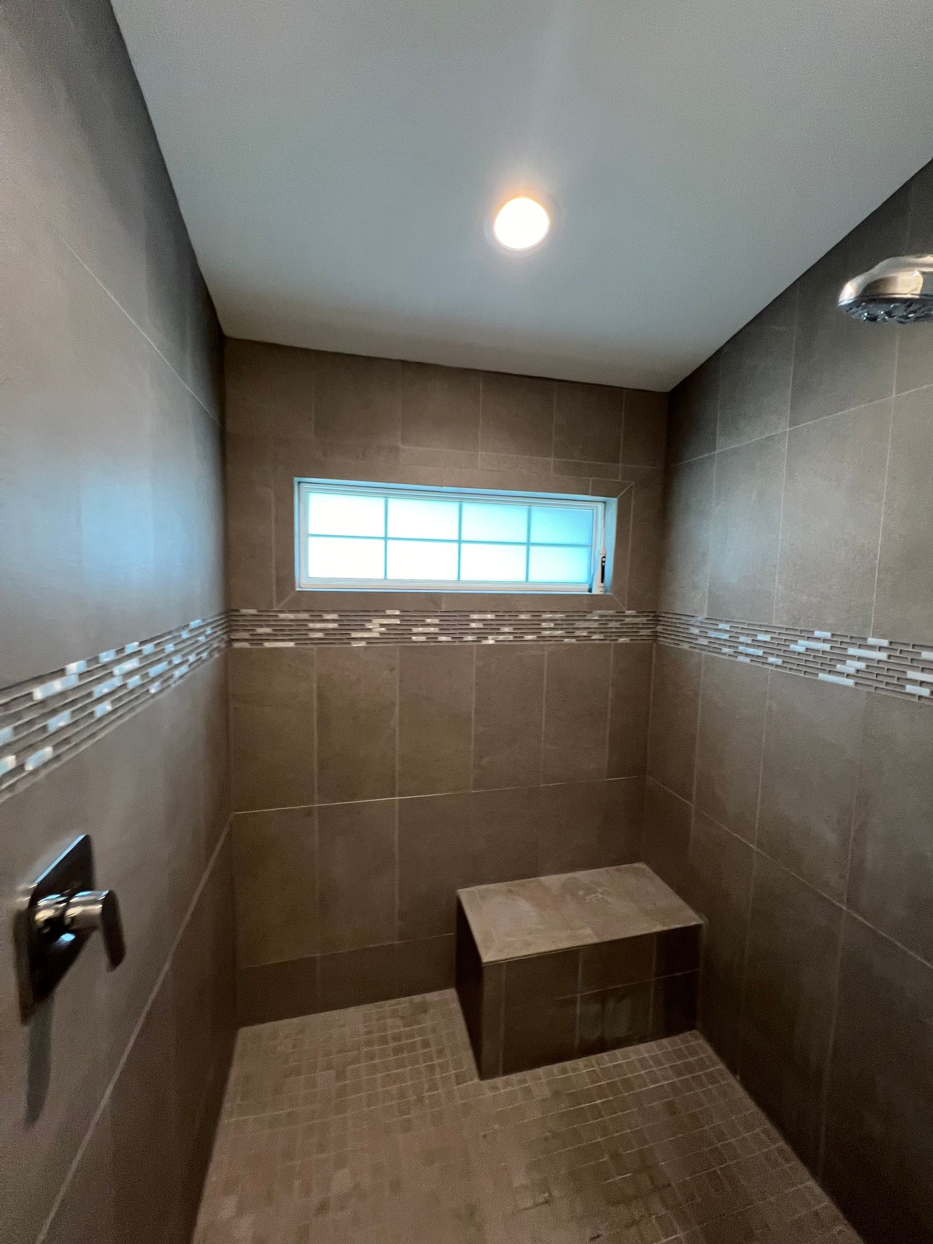 Shower with brown tile walls, small window, bench, and chrome fixtures.