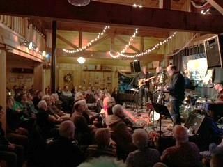Band performing on stage in a warmly lit venue. Audience watches, seated. String lights overhead.