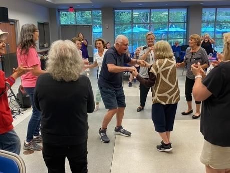 People dancing in a community center, some clapping. Indoors, well-lit, with a pool visible through windows.
