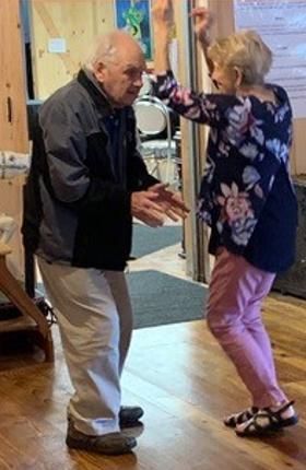 An elderly couple dancing indoors. The woman has her arms raised.