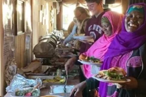People serving food from a buffet table; smiling woman in hijab holds a plate.