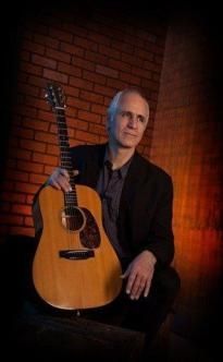 Man holding acoustic guitar, set against a brick wall.