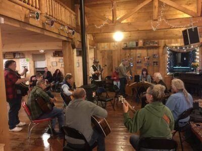 People playing music in a warmly lit lodge. Musicians play guitars, banjo, and other instruments. Audience watches.