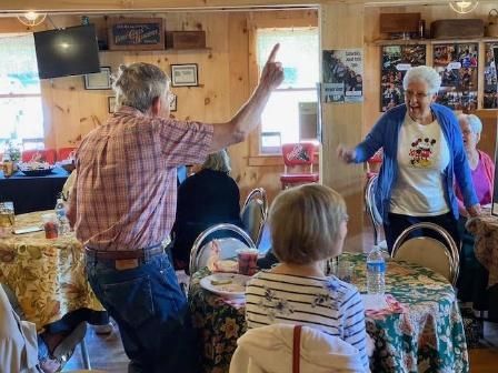 People dancing at a gathering. One man points up, a woman smiles and dances. Tables set in a wood-paneled room.